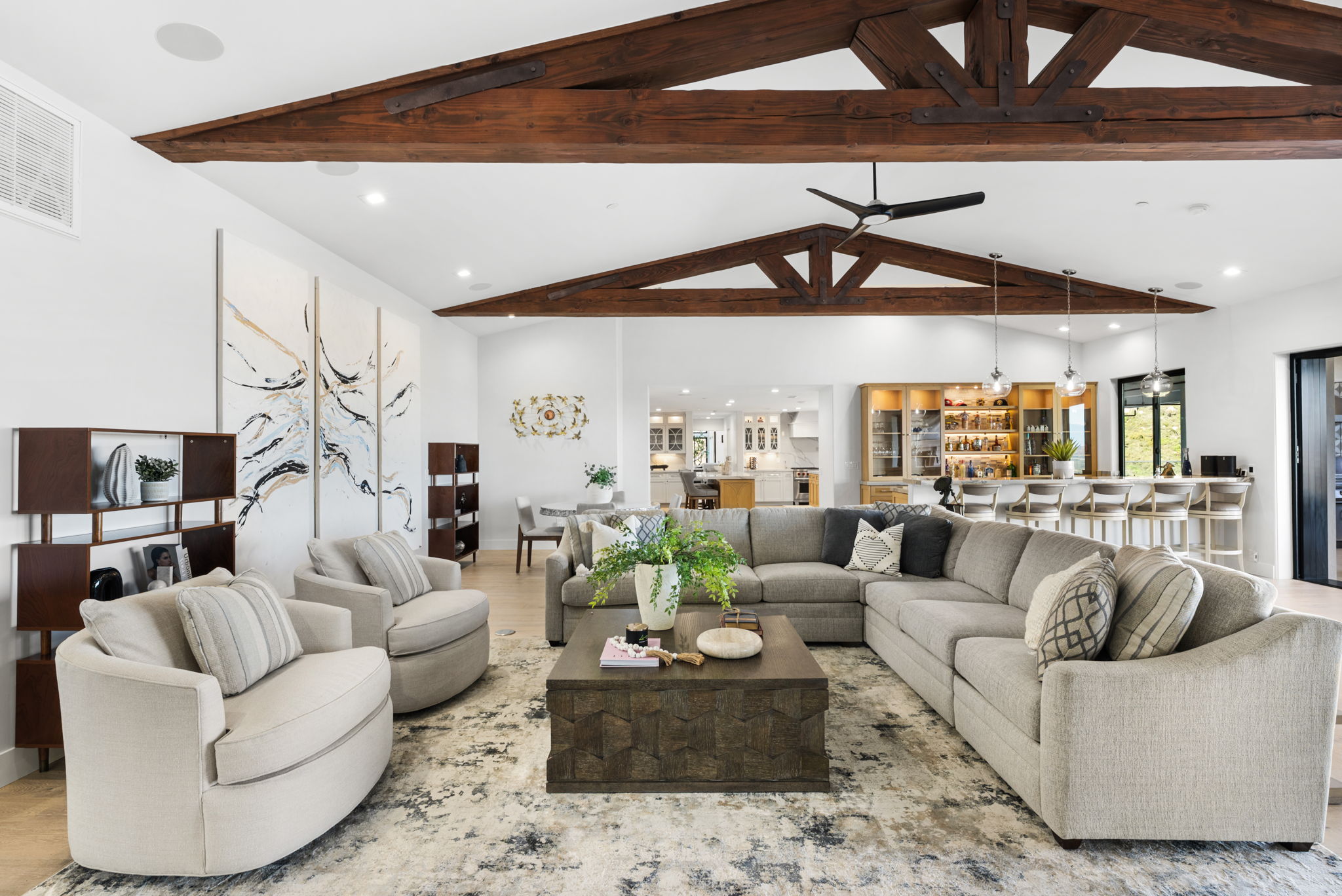 This spacious, open-concept living area features stunning vaulted ceilings with exposed wood trusses and a modern fan. The room is anchored by a large, comfortable gray sectional sofa and a dark wood coffee table, set against a backdrop of a neutral, textured rug and contemporary abstract art. With a seamless transition to the kitchen and bar area in the background, this space is designed for both relaxation and high-end entertaining.