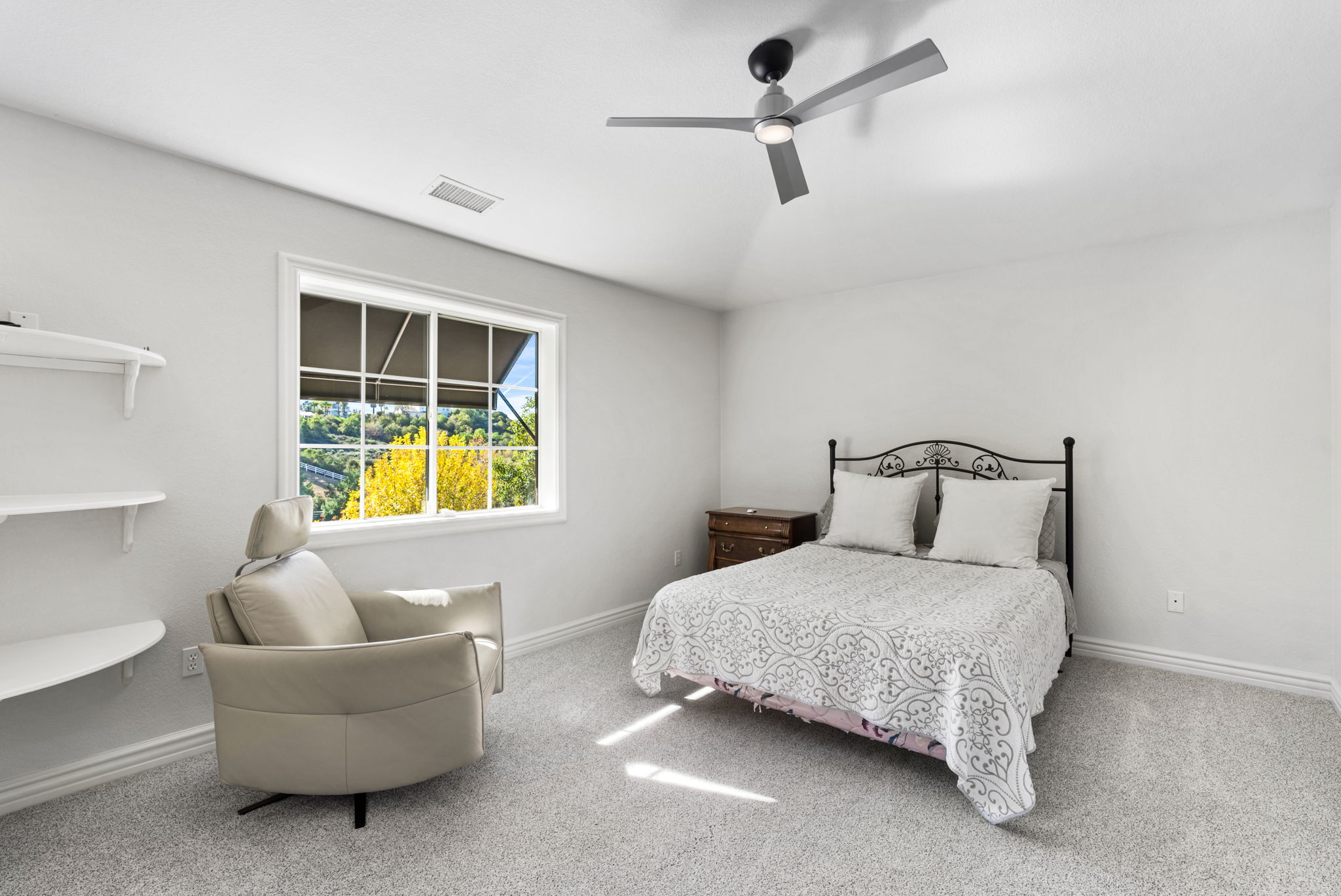 This bright and airy bedroom features a wrought-iron bed frame with a patterned quilt, a comfortable modern lounge chair, and built-in wall shelving. The room is enhanced by neutral carpeting and a large window that allows for natural light while offering views of the surrounding landscape. The simple, clean aesthetic creates a relaxing and versatile living space.