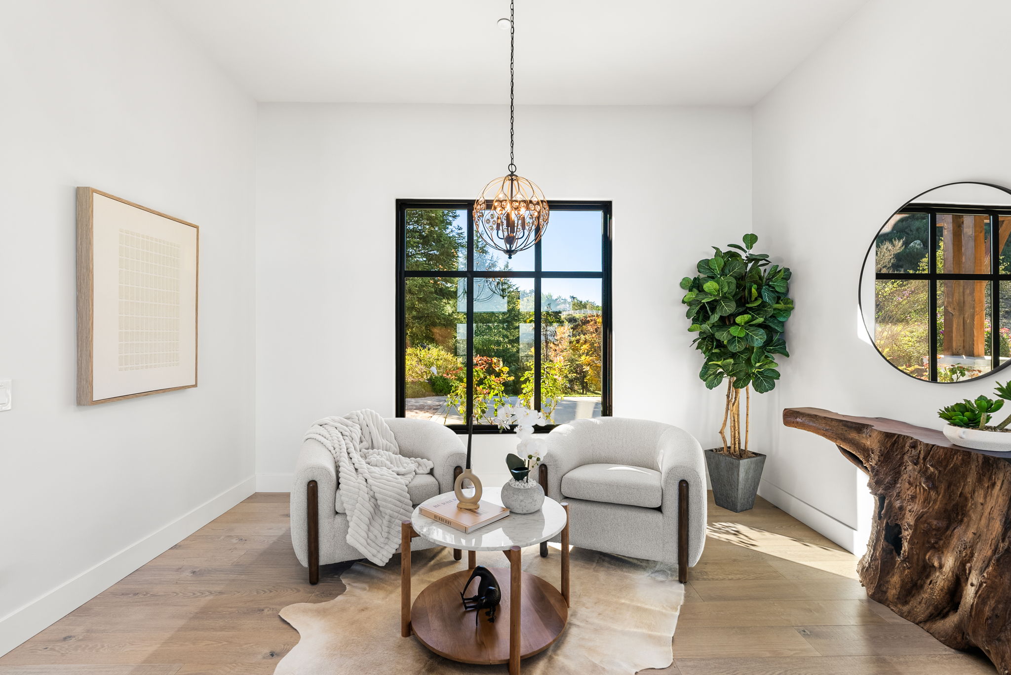This bright and elegant sitting area features a pair of plush, light-colored armchairs centered around a round marble-topped coffee table. A floor-to-ceiling window provides a picturesque view of the outdoors, while a statement pendant light and unique wood-carved console table add sophisticated character to the room. The space is finished with high-quality hardwood floors, a neutral color palette, and tasteful decorative accents, creating a warm and inviting atmosphere.