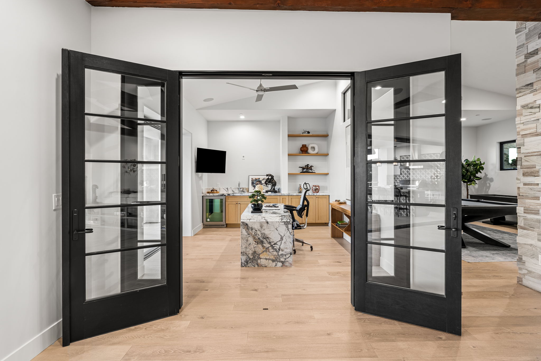 This modern home office is elegantly framed by striking black-trimmed French doors, revealing a luxurious marble-topped desk and high-end cabinetry. The space features light hardwood floors, sleek open shelving, and integrated office amenities, including a wine cooler and wall-mounted television. The clean, contemporary aesthetic, highlighted by vaulted ceilings and a ceiling fan, creates an inviting and professional environment.