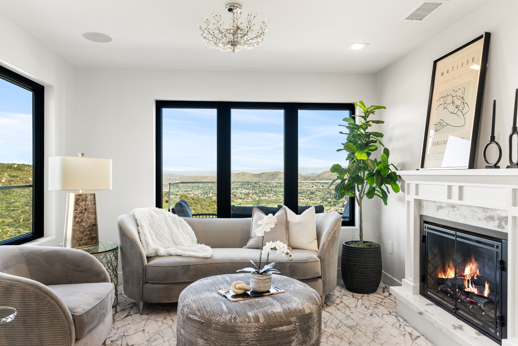This elegant living room features a cozy seating area centered around a fireplace and a large picture window with stunning panoramic mountain and valley views. The space is decorated with a sophisticated neutral color palette, including a plush grey sofa, a matching round ottoman, and contemporary art, creating a refined and relaxing atmosphere. High-end touches like the crystal chandelier and high-quality finishes make this room a luxurious highlight of the home.