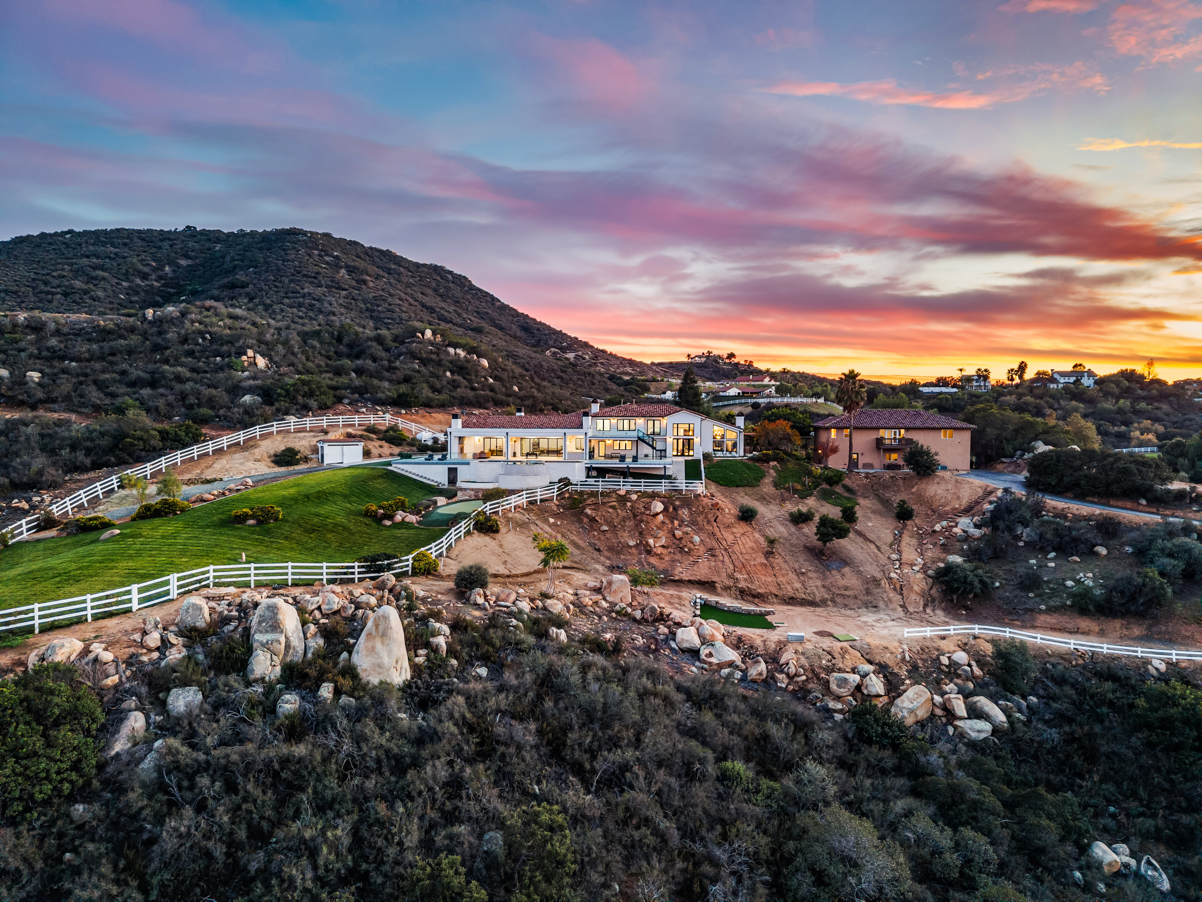 This stunning aerial view captures a sprawling, modern-style estate perched on a hillside at sunset, surrounded by natural rocky terrain and native landscaping. The property features a pristine, manicured green lawn, a white-fenced perimeter, and a contemporary home with expansive windows that glow warmly in the twilight. The dramatic mountain backdrop and vibrant, colorful sky create a luxurious and serene atmosphere highly desirable for premium real estate.