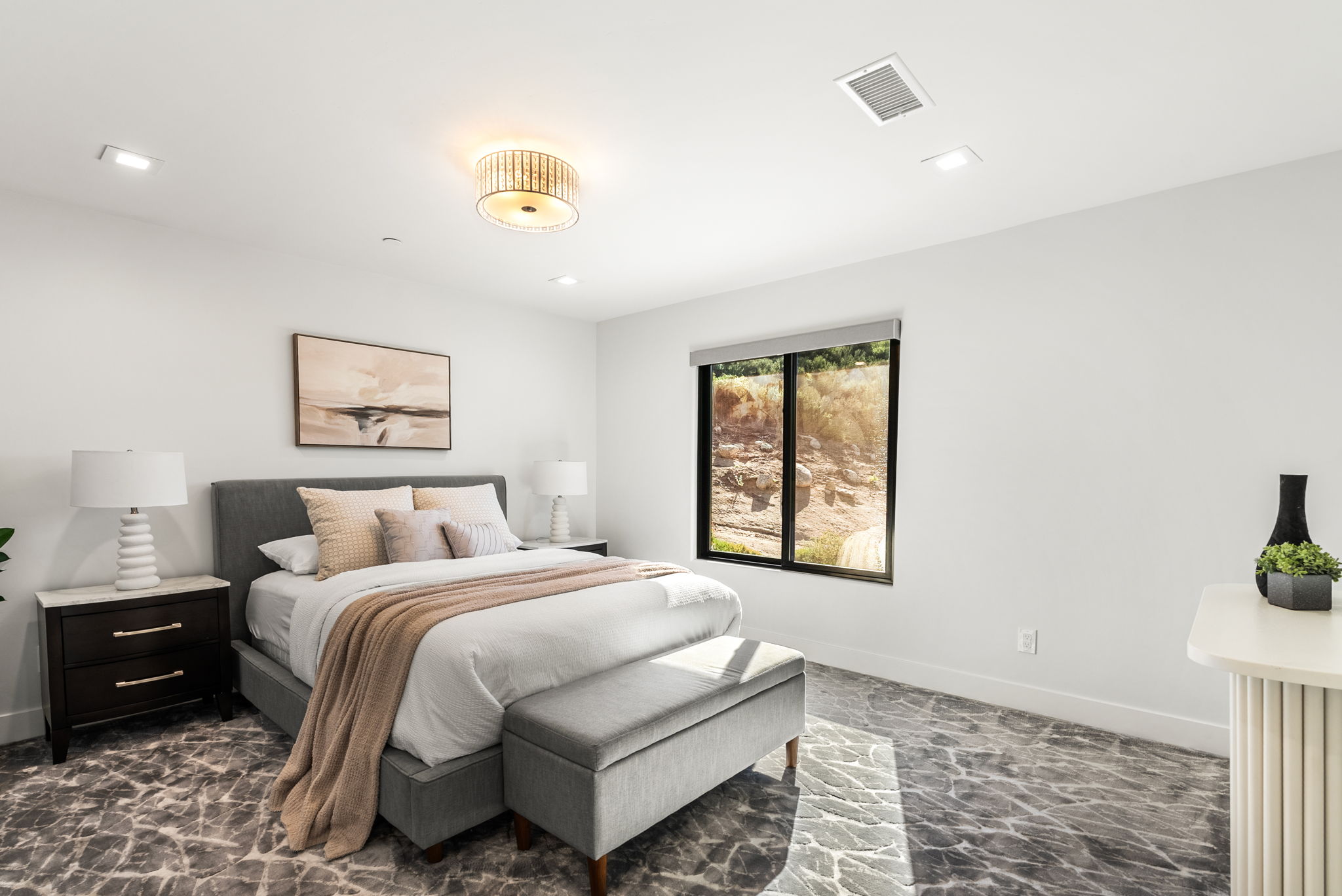 This modern primary bedroom features a neutral color palette, characterized by a tufted gray bed frame, soft beige bedding, and a plush patterned carpet. Large windows provide natural light and a view of the natural landscape, while a sleek bedside table and decorative accents contribute to a clean, contemporary aesthetic. The room is well-lit with a stylish chandelier and recessed lighting, creating a calm and inviting atmosphere.