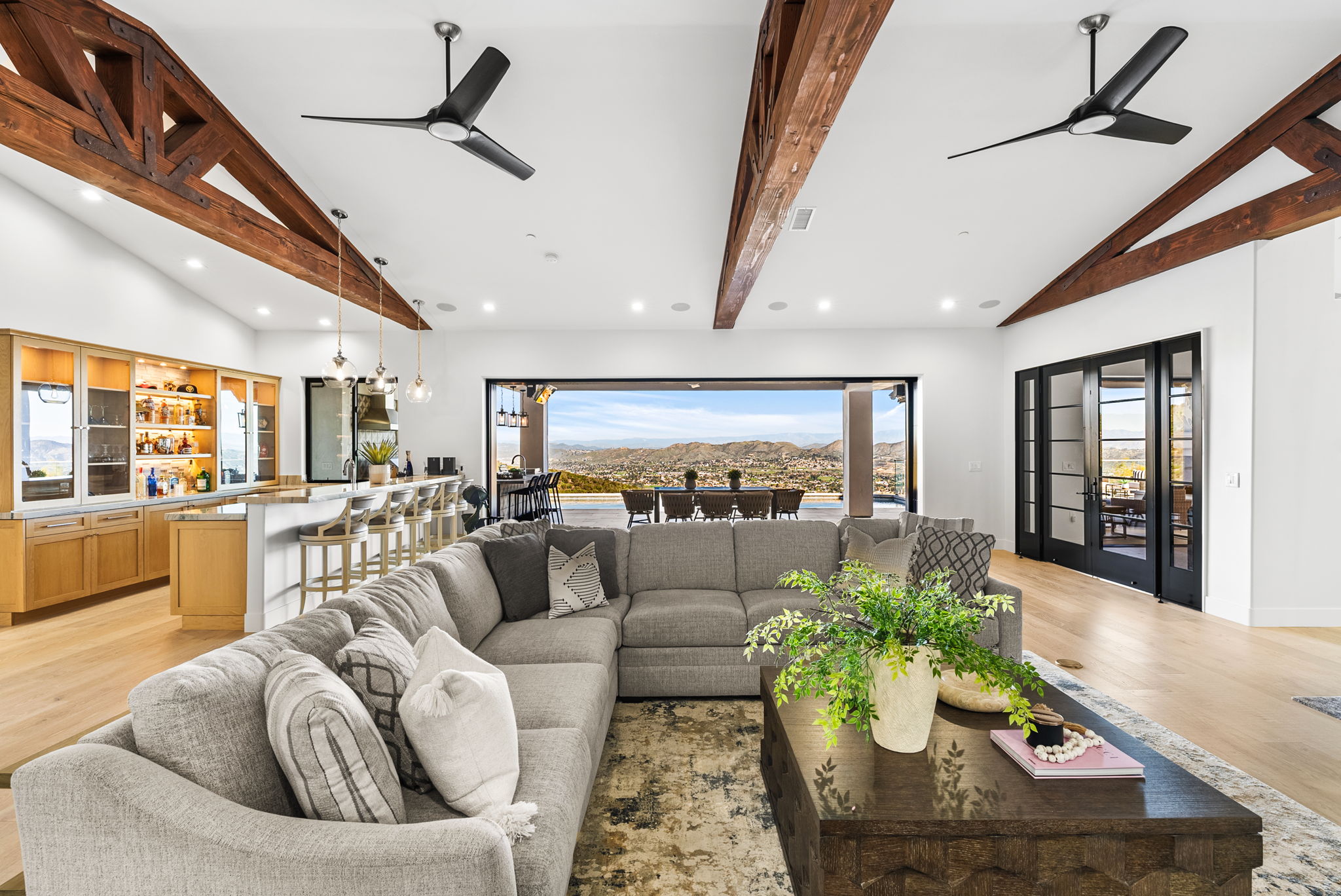 This expansive, open-concept living area features a large, plush sectional sofa and a dark wood coffee table as the centerpiece, perfect for relaxed entertaining. The space is defined by dramatic vaulted ceilings with exposed wood beams and a seamless indoor-outdoor transition via expansive sliding glass doors that reveal stunning panoramic mountain views. A sophisticated kitchen with a breakfast bar and warm wood cabinetry sits adjacent to the living space, creating a cohesive and luxurious home environment.
