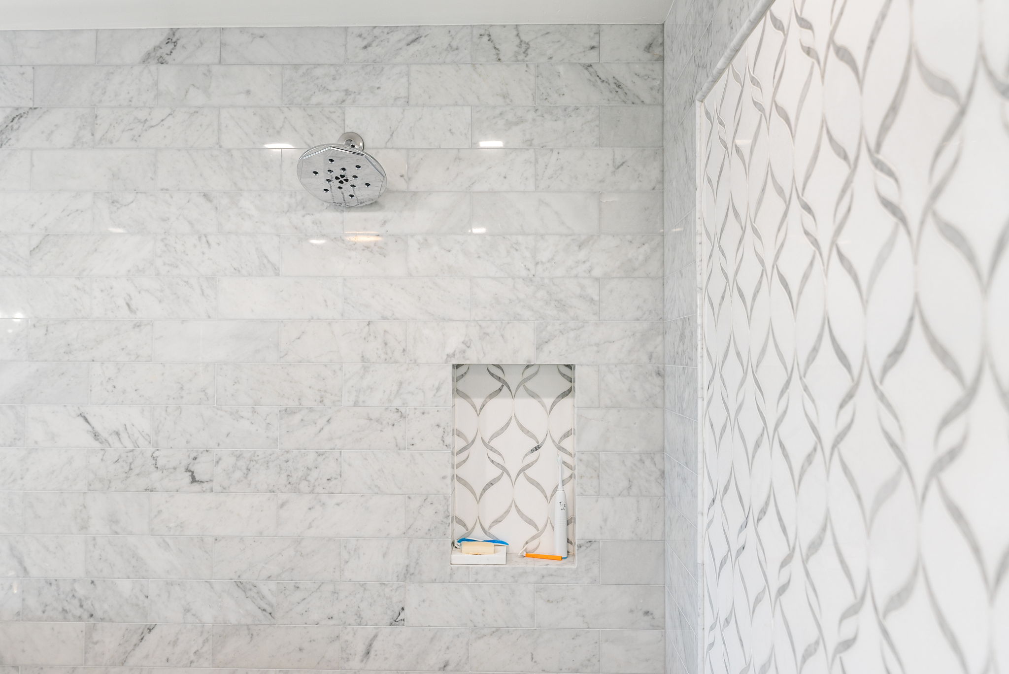 This shower area features elegant white marble subway-style wall tiles paired with a striking patterned accent tile inside the recessed niche and on the adjacent wall. The space is equipped with a chrome shower head and provides functional storage within the built-in alcove, creating a luxurious and cohesive aesthetic.