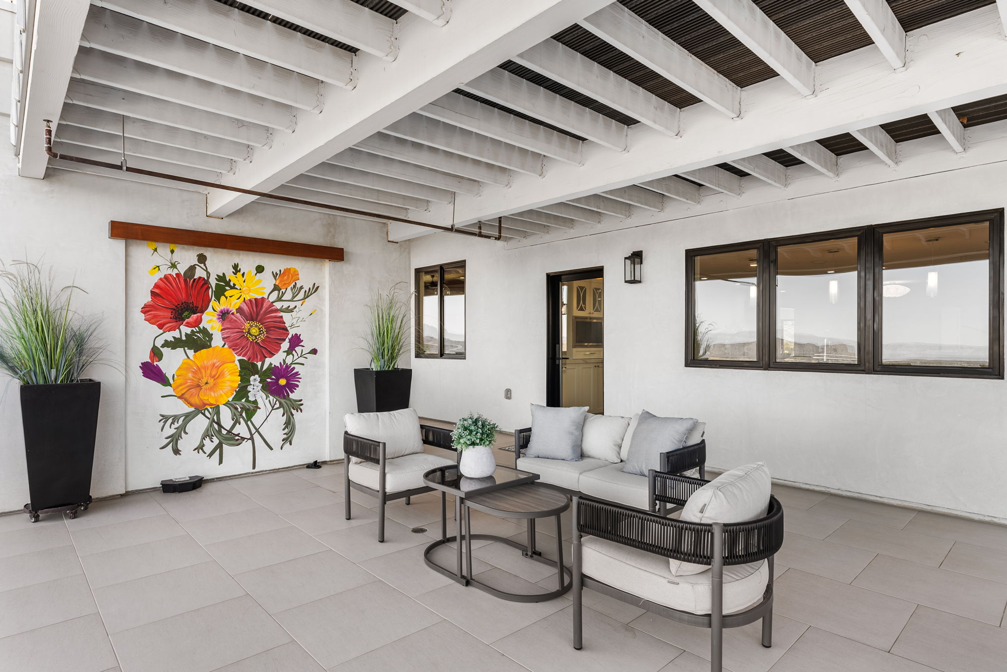 This covered patio offers a sophisticated outdoor living space featuring a comfortable seating arrangement with modern, neutral-toned furniture. The area is highlighted by a vibrant, artistic floral mural on the wall and sleek tile flooring, creating a welcoming and stylish environment. The combination of the open-air design, tasteful decor, and well-maintained finishes makes this an ideal space for relaxation and entertaining.