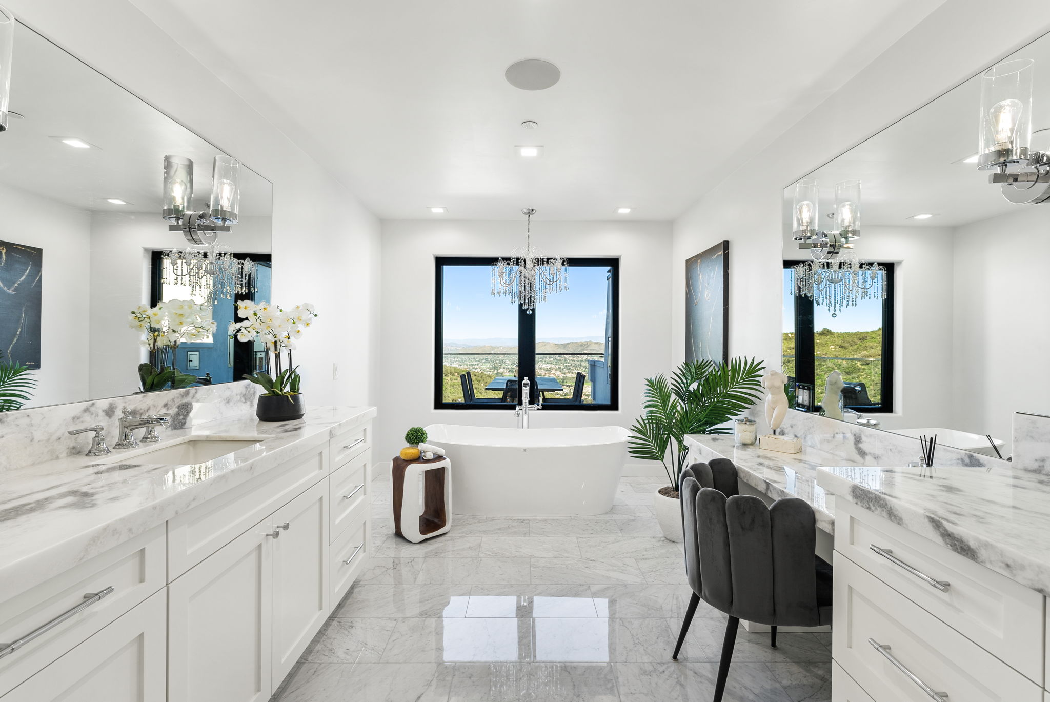 This luxurious primary bathroom features a freestanding soaking tub centered in front of a large window with panoramic views. The space is elegantly finished with white marble countertops, dual vanities, and a built-in makeup vanity with a velvet chair. Sparkling crystal chandeliers and high-end finishes create a sophisticated and bright atmosphere.
