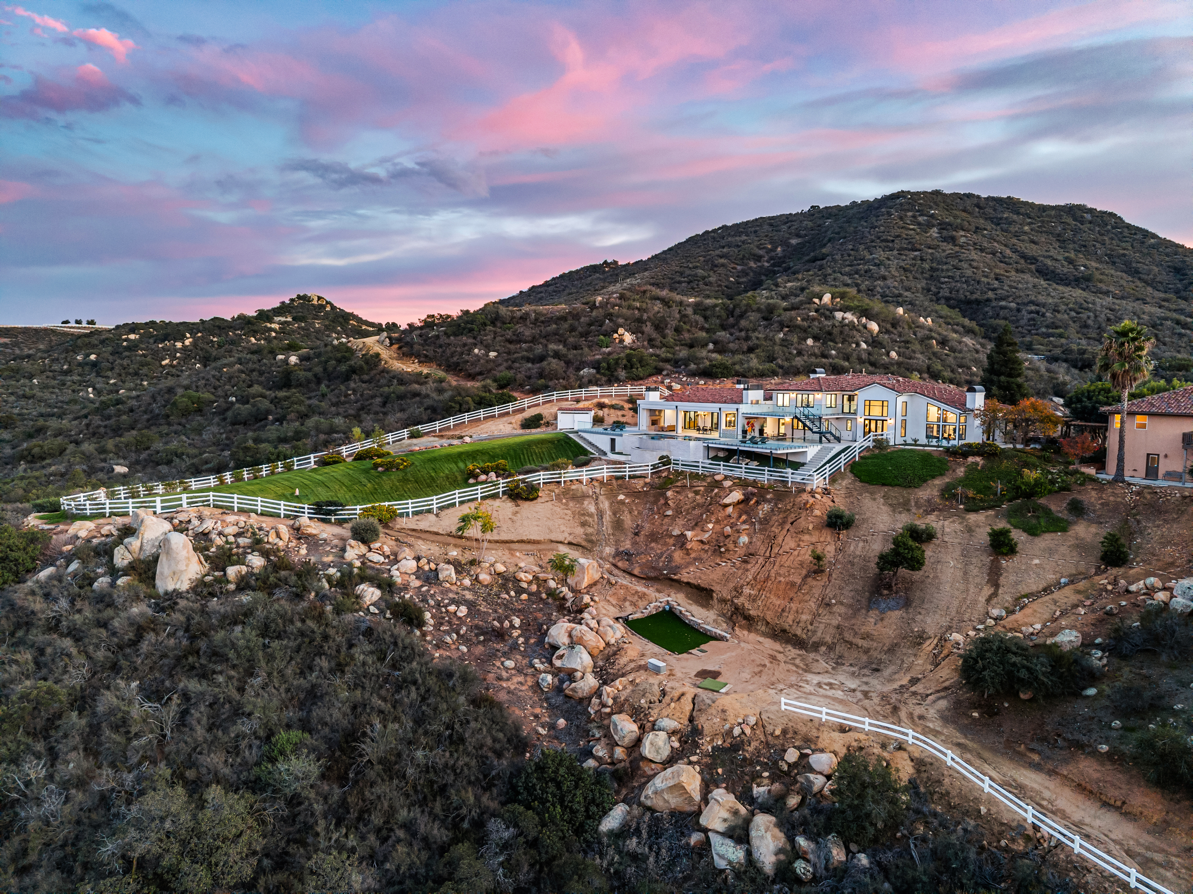 This stunning aerial view captures a sprawling multi-level residential estate nestled against a rugged hillside at dusk. The property features expansive manicured lawns, a tiered patio with a pool, and white post-and-rail fencing, all illuminated by warm house lighting under a vibrant pink and purple sunset sky. The architectural style blends modern elements with traditional hillside landscape integration, offering a commanding presence in a natural, secluded setting.