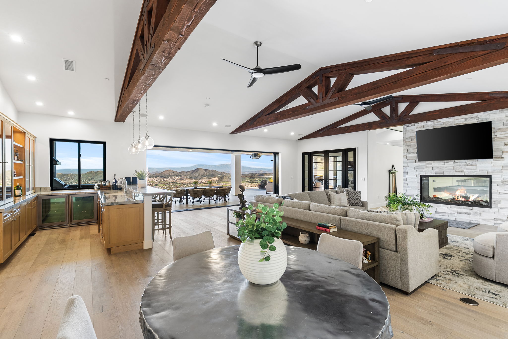 This spacious open-concept living area features stunning exposed wood trusses, light hardwood flooring, and expansive glass sliding doors that reveal panoramic mountain views. The room is centered around a sleek stone fireplace with an integrated television, complemented by a comfortable sectional and a modern dining area. The blend of rustic architectural elements and contemporary design creates an airy, luxurious atmosphere ideal for indoor-outdoor living.