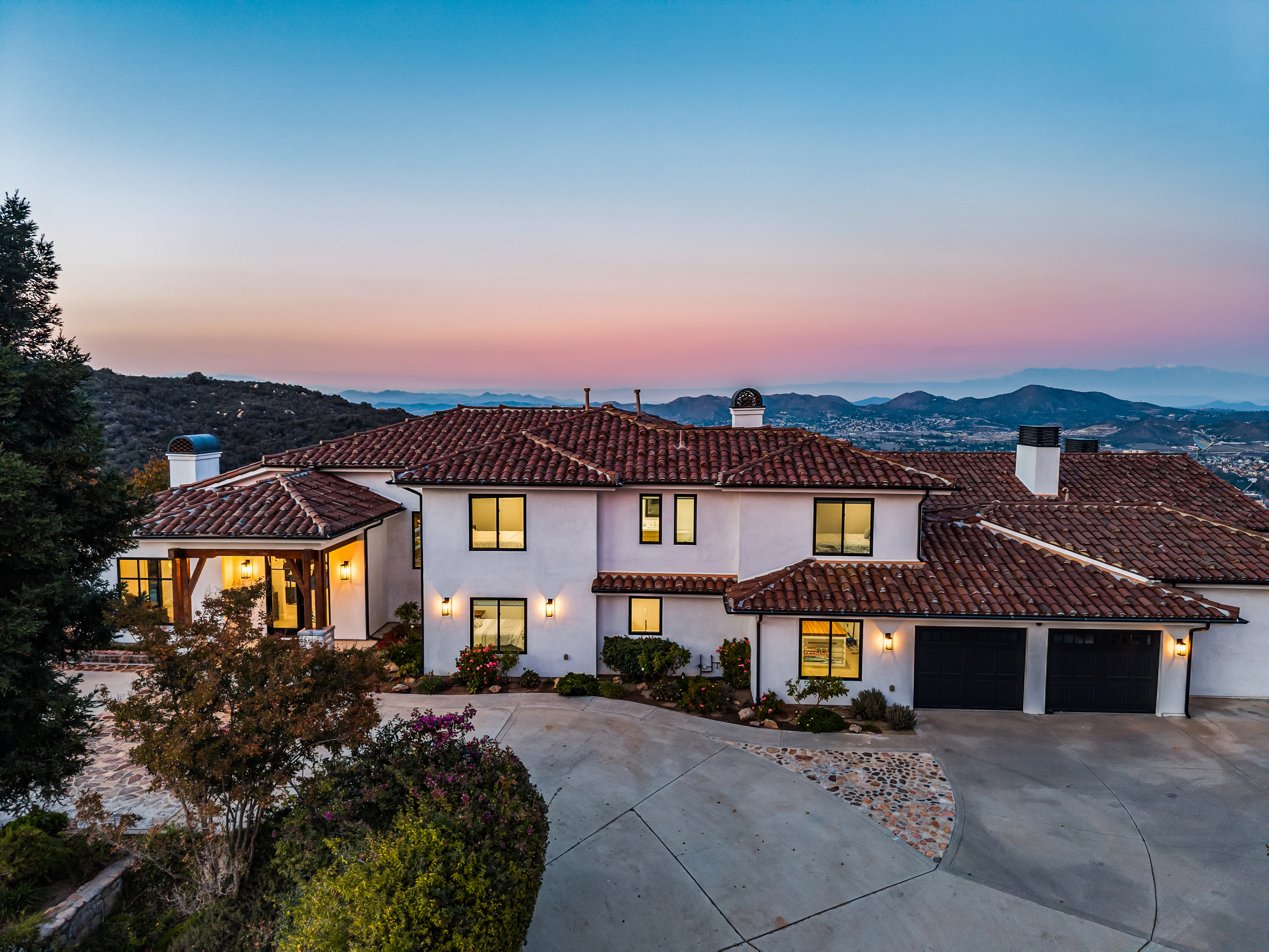 This stunning Mediterranean-style residence is captured from an elevated aerial perspective at dusk, showcasing its expansive tile roof, crisp white exterior, and beautiful landscape integration. The property features a spacious multi-car garage, large windows glowing with warm interior light, and a grand driveway that winds through the meticulously maintained front grounds. Its commanding position overlooking distant hills and valley views creates a sophisticated and desirable suburban retreat.