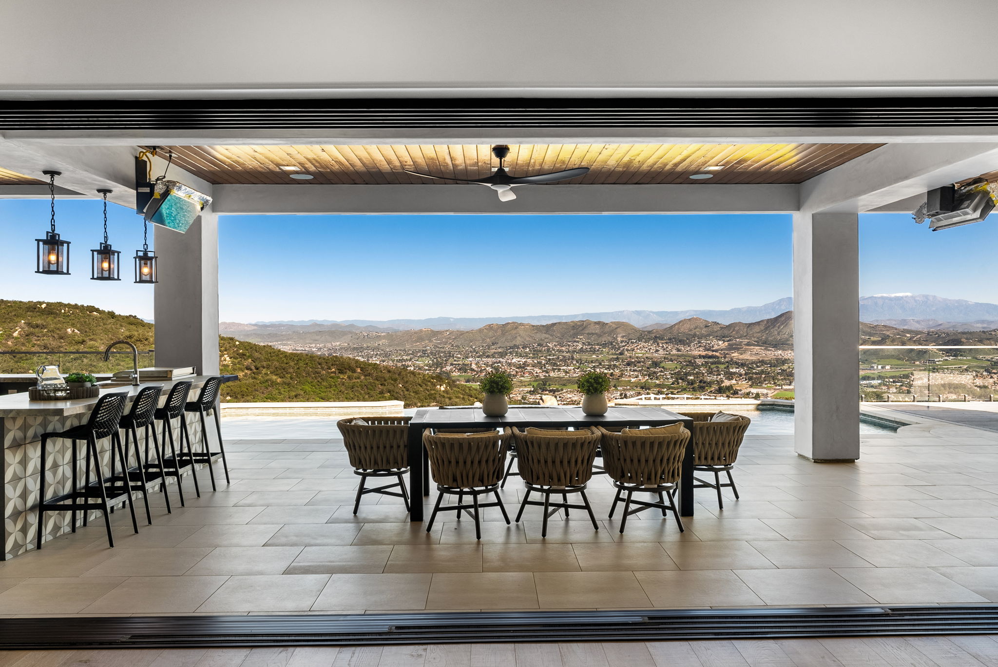 This luxurious outdoor living space features a covered patio that blends seamlessly with the scenic valley views through an expansive open-air design. The area is equipped with a modern bar with black mesh stools and a spacious dining set, making it perfect for indoor-outdoor entertaining. The clean, contemporary aesthetic is enhanced by recessed lighting, a ceiling fan, and high-end finishes, offering an inviting and sophisticated atmosphere.