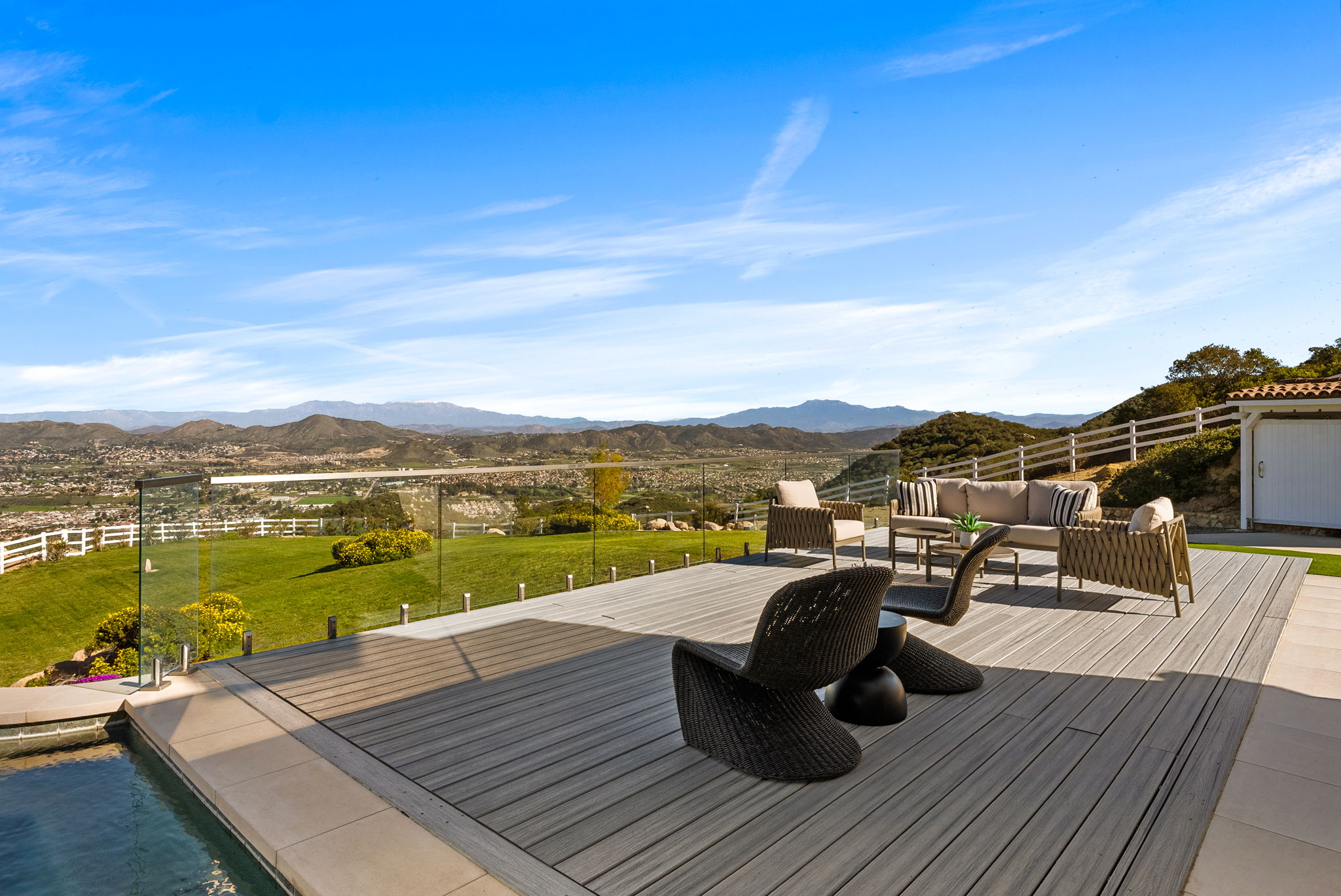 This stunning outdoor deck offers a luxurious living space with panoramic mountain and valley views. The area features premium modern wicker patio furniture, including a sofa set and lounge chairs, arranged on high-quality composite decking. A sleek glass railing preserves the unobstructed scenic vista, creating an ideal setting for outdoor entertaining.
