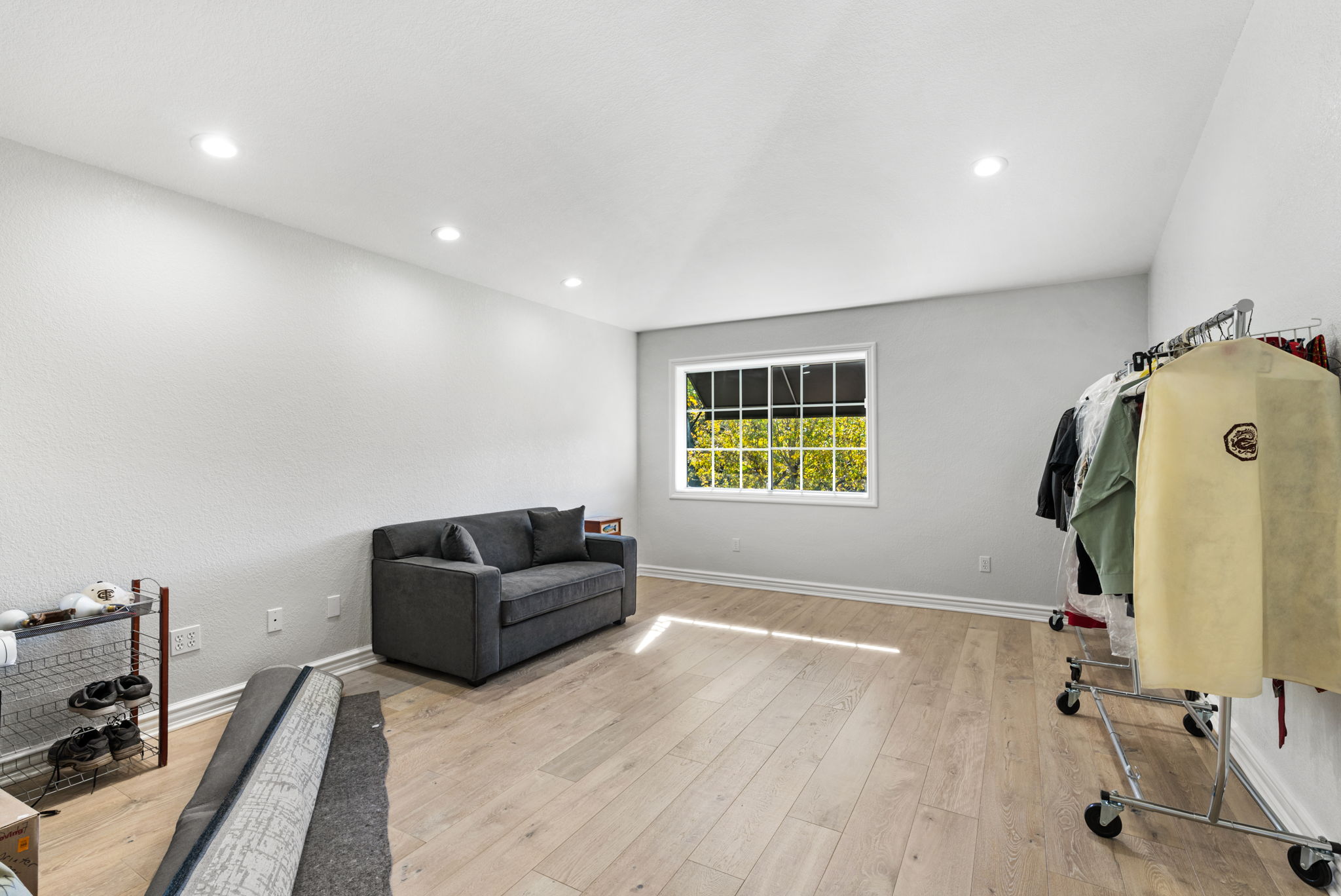 This room features light-colored laminate flooring, neutral white walls, and recessed lighting, creating a bright and clean atmosphere. It is furnished with a dark gray sofa, a wire shoe storage unit, and a rolling garment rack, making it a versatile space that could serve as a bedroom or multi-purpose area. A single window provides natural light and a view of the greenery outside.