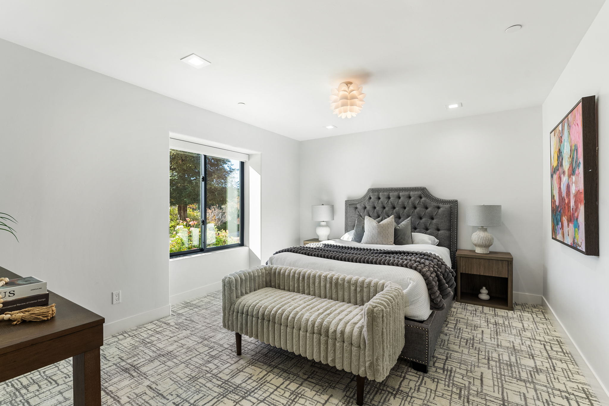 This modern bedroom features a tufted grey upholstered bed frame, a textured bench, and a sophisticated color palette of whites, greys, and warm wood tones. An abstract art piece hangs on the wall, complementing the contemporary decor, while a large window invites natural light into the space. The room is finished with patterned carpeting and elegant lighting, creating a comfortable and inviting atmosphere.