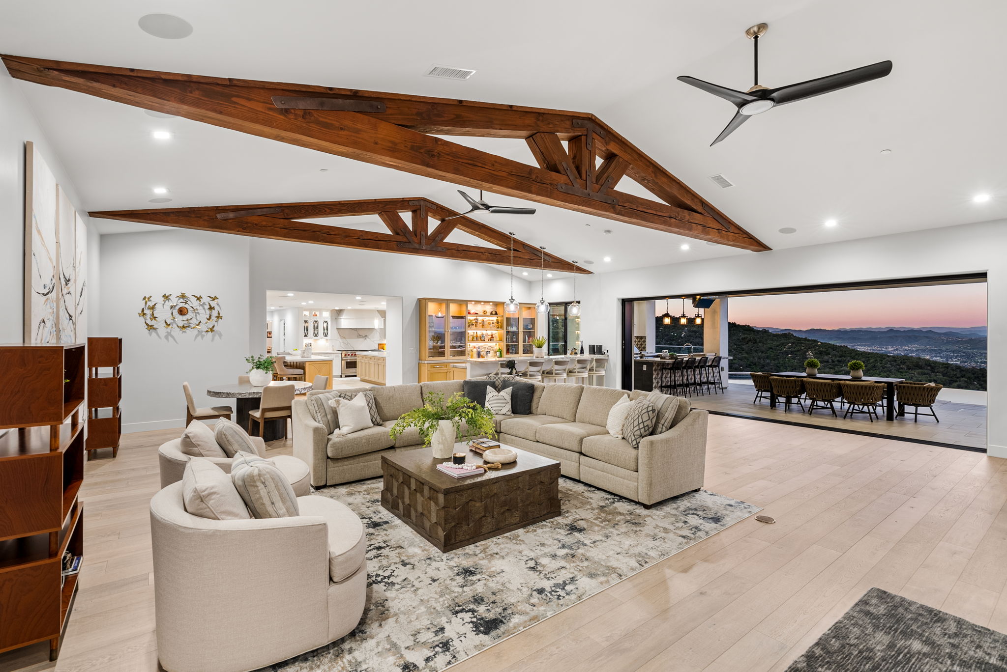 This spacious, open-concept living area features stunning exposed wooden vaulted ceilings and a seamless transition to an expansive outdoor deck with panoramic hillside views. The room is anchored by a large, comfortable neutral-toned sectional sofa, a textured area rug, and a modern wooden coffee table, all set beneath high-end ceiling fans. A wet bar with pendant lighting and a partial view of the adjacent kitchen make this an ideal space for entertaining.