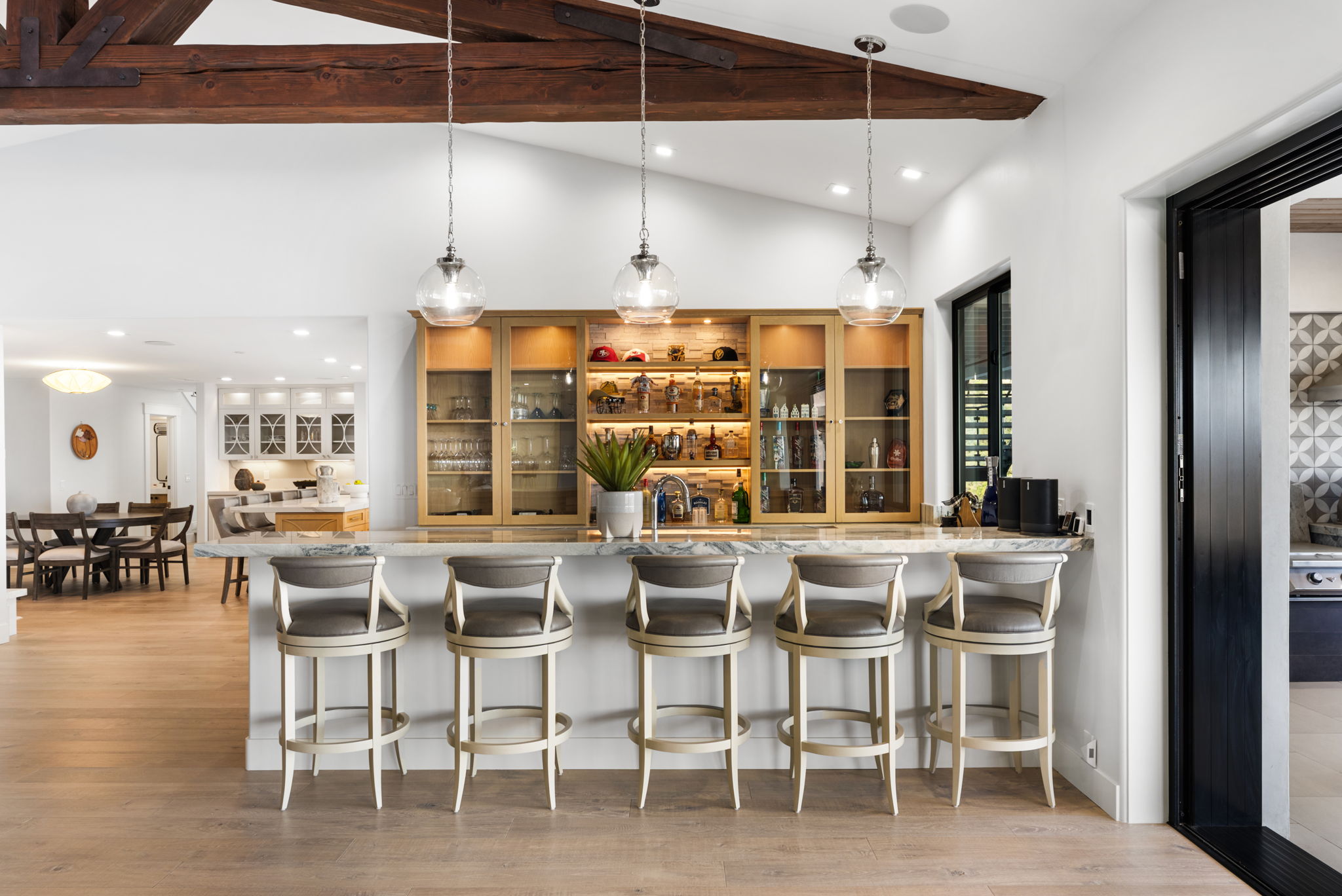 This interior space features a sophisticated wet bar area with custom light wood cabinetry, illuminated glass shelving, and a marble countertop with five bar stools. The room boasts a high, vaulted ceiling with exposed wooden beams and stylish glass pendant lights, creating an inviting and luxurious atmosphere. Seamlessly integrated into the open floor plan, it offers a perfect transition between the dining area and the main living space.