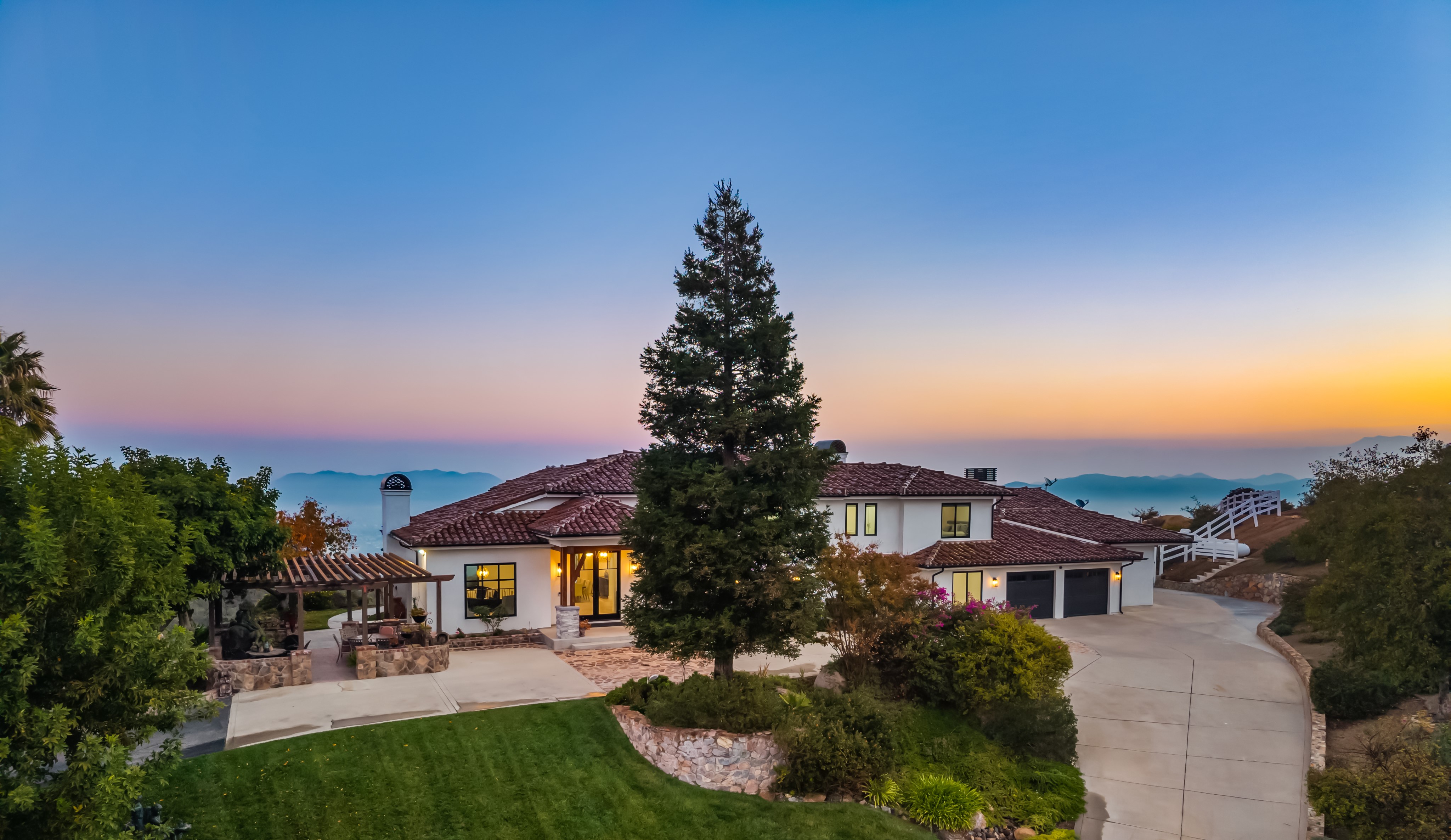 This stunning Mediterranean-style estate is captured from an elevated perspective, showcasing its sprawling layout and scenic hillside location. The property features a white exterior, a classic clay tile roof, and a meticulously landscaped front yard with a prominent mature tree as a focal point. A paved driveway leads to a multi-car garage, while a stone-accented patio area offers an inviting space for outdoor dining, all set against a breathtaking sunset backdrop.
