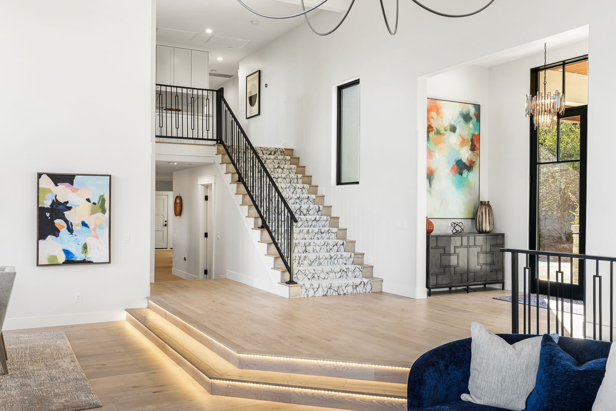 This modern, light-filled foyer features an elegant staircase with a patterned runner and wrought-iron railings. A multi-level landing with integrated step lighting leads into the space, which is accented by contemporary artwork, a sleek dark sideboard, and high-end finishes. The open-concept design creates a sophisticated and airy transition between the different levels of the home.