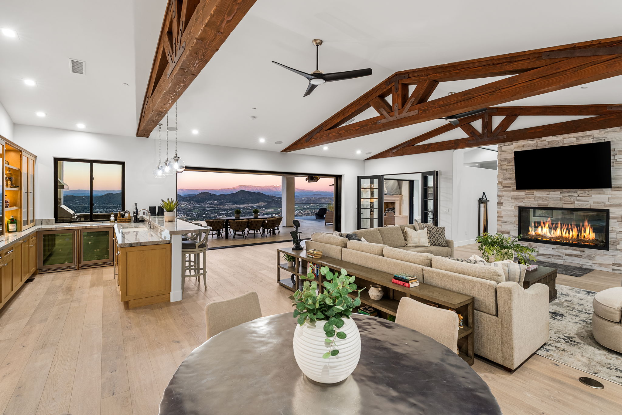 This spacious, open-concept living area features stunning vaulted ceilings with exposed wood trusses and a modern fireplace with stone cladding. A large glass wall slides open to a seamless indoor-outdoor transition, revealing a panoramic view of the mountains. The room is anchored by a comfortable, neutral-toned sectional sofa and flows effortlessly into a well-appointed kitchen and dining space.