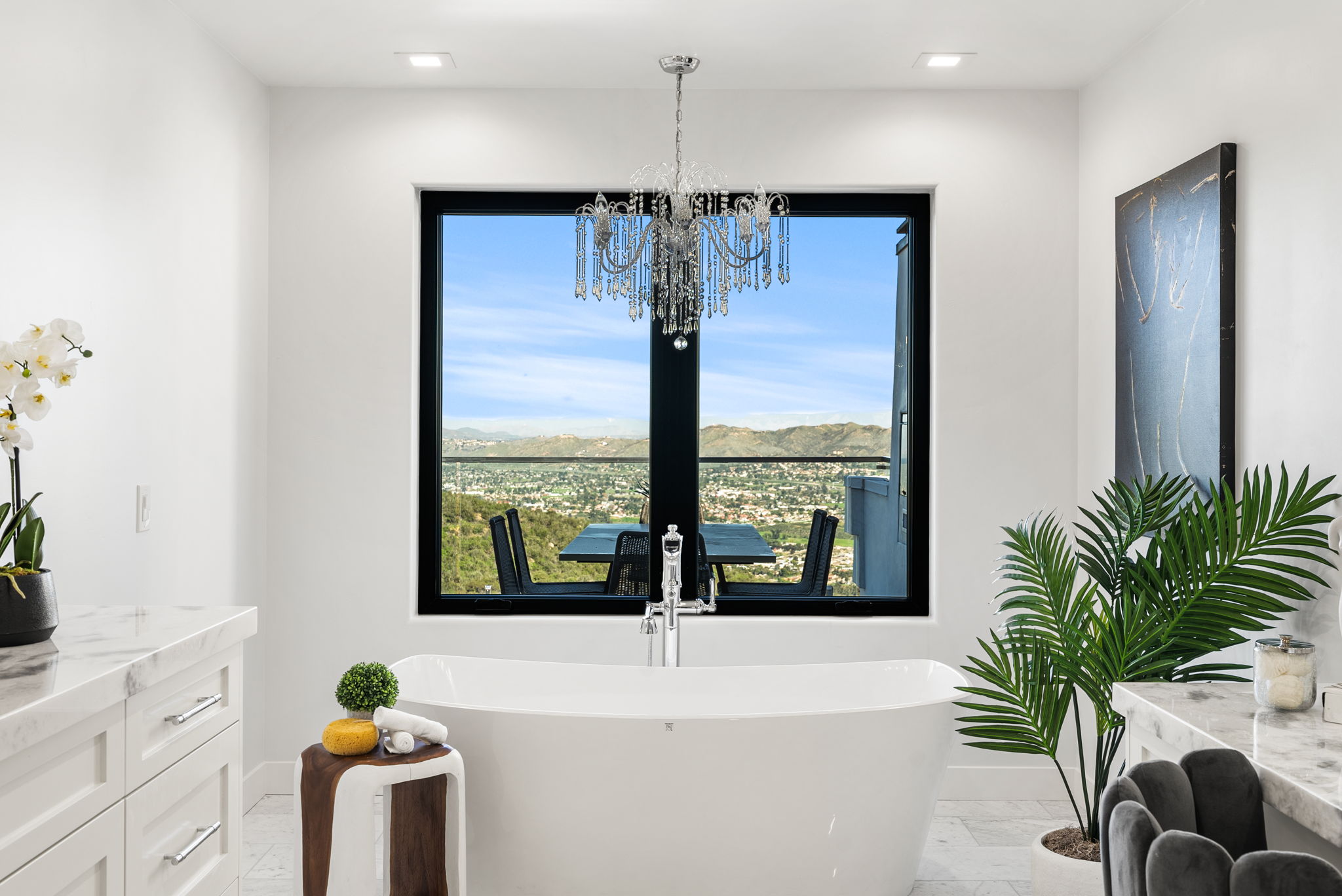 This modern, luxurious primary bathroom features a standalone soaking tub centered beneath a large window that frames stunning panoramic mountain and valley views. A glamorous crystal chandelier hangs above the tub, complemented by clean white cabinetry, marble countertops, and vibrant indoor plants that add a touch of freshness. The overall aesthetic is serene and sophisticated, creating a spa-like atmosphere perfect for relaxation.