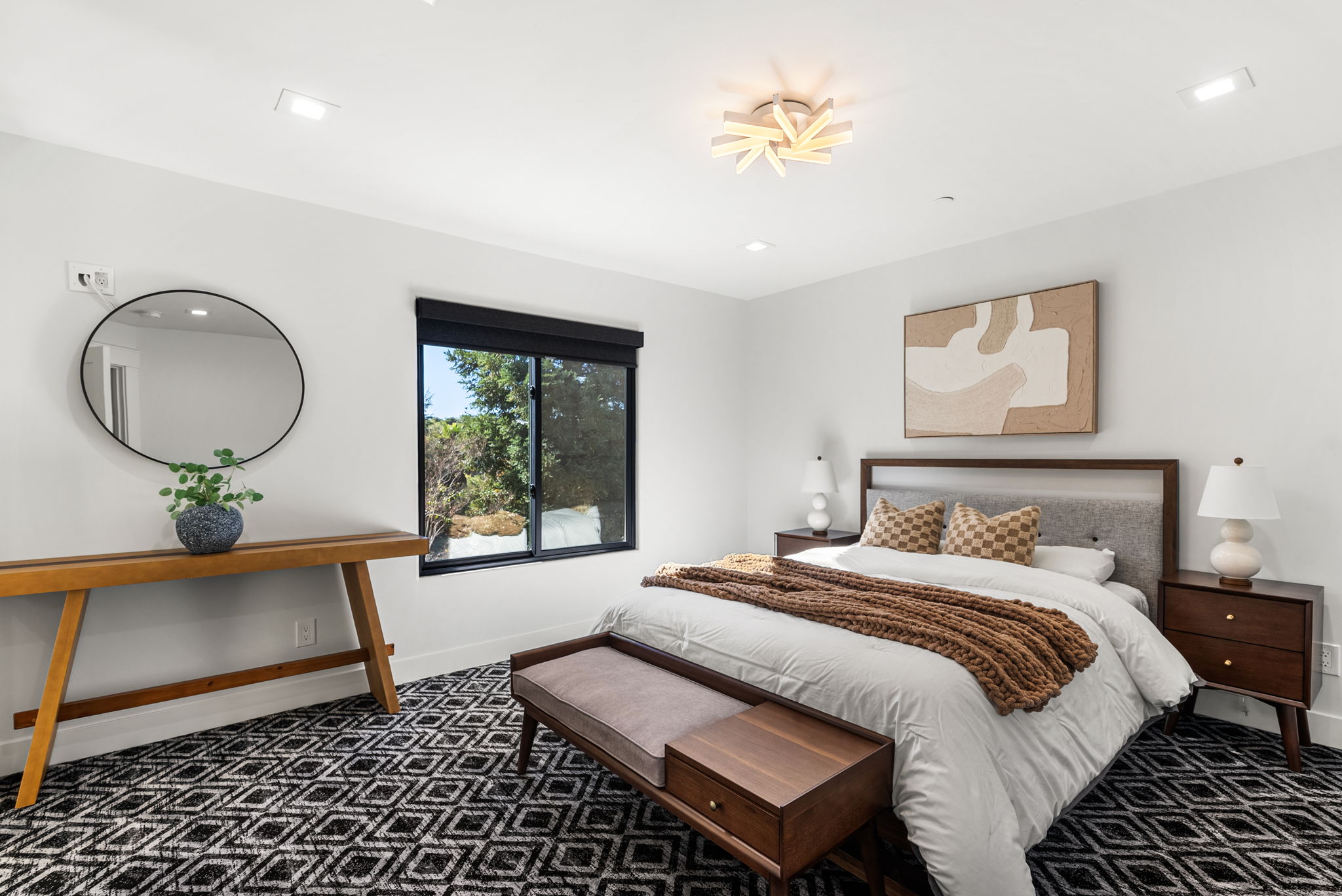 This primary bedroom features a modern, clean aesthetic with a comfortable bed styled with a chunky knit throw and patterned pillows. The space is highlighted by a distinctive geometric patterned carpet, mid-century modern bedside tables, and a sleek wooden console table paired with a circular mirror. A large window provides natural light and a pleasant outdoor view, creating a bright and inviting atmosphere.