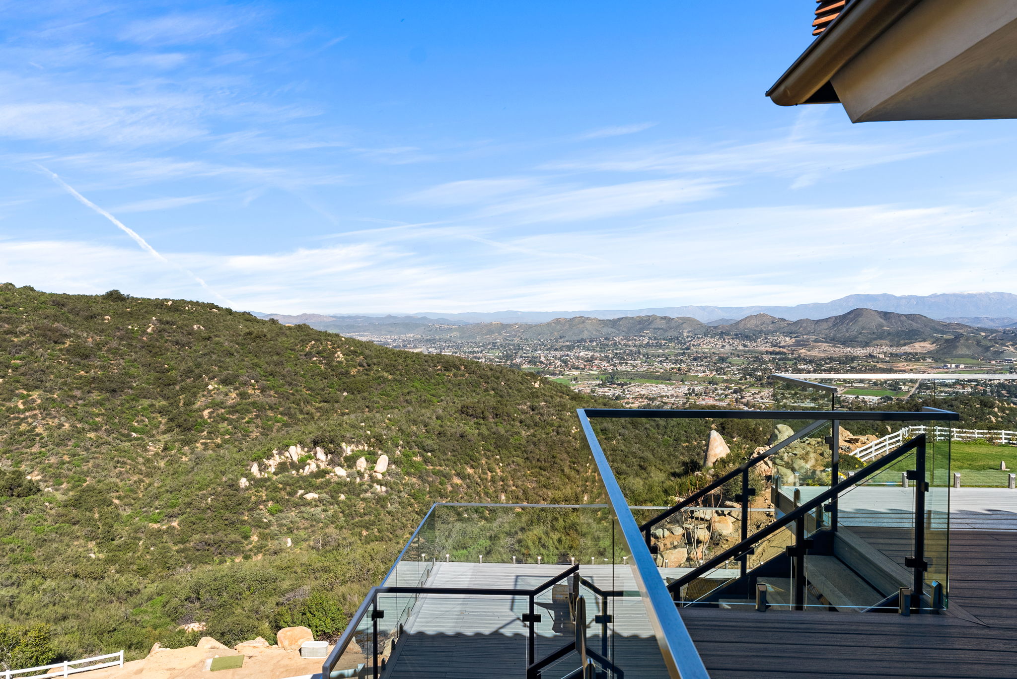 This image showcases a stunning multi-level outdoor deck featuring modern glass railings that provide an unobstructed, panoramic view of the rolling hills and the valley town below. The sleek, tiered architectural design enhances the feeling of luxury, seamlessly blending the home's living space with the expansive natural scenery. It creates an ideal setting for outdoor entertaining or quiet relaxation while enjoying the sweeping vista.