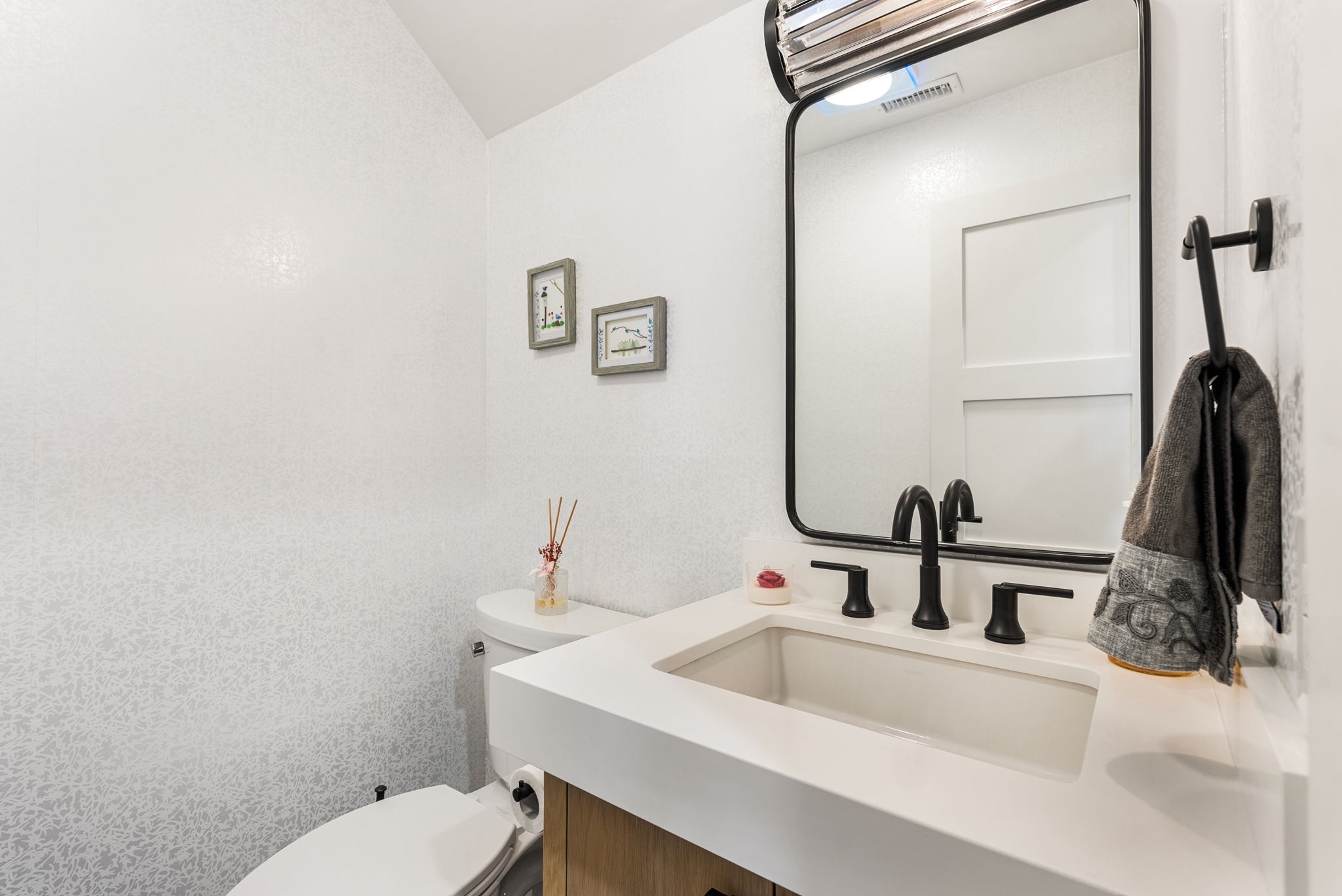 This compact powder room features modern design elements, including a vanity with a sleek white countertop and matte black fixtures. The space is accented by patterned wallpaper, a black-framed mirror, and framed wall art, creating a clean and contemporary aesthetic. A toilet is positioned conveniently adjacent to the vanity, optimizing the small floor plan.