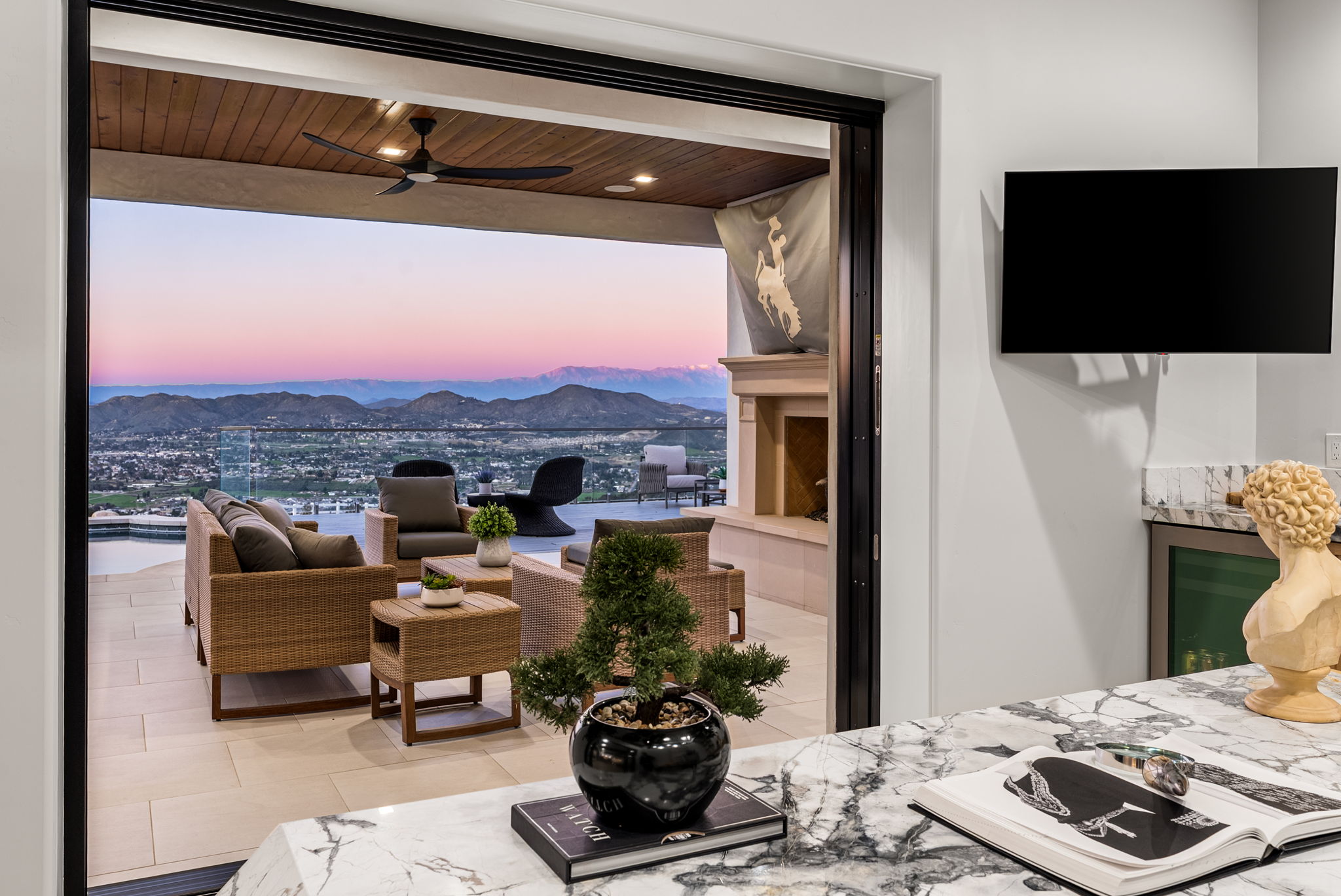 This image showcases a luxurious indoor-outdoor living space, featuring a seamless transition from a marble-topped interior bar to a spacious covered patio. The outdoor area is furnished with high-end wicker lounge seating and a fireplace, all set against a stunning panoramic view of mountains and a sunset sky. The combination of elegant interior decor and expansive, scenic outdoor living makes this home ideal for sophisticated entertaining.