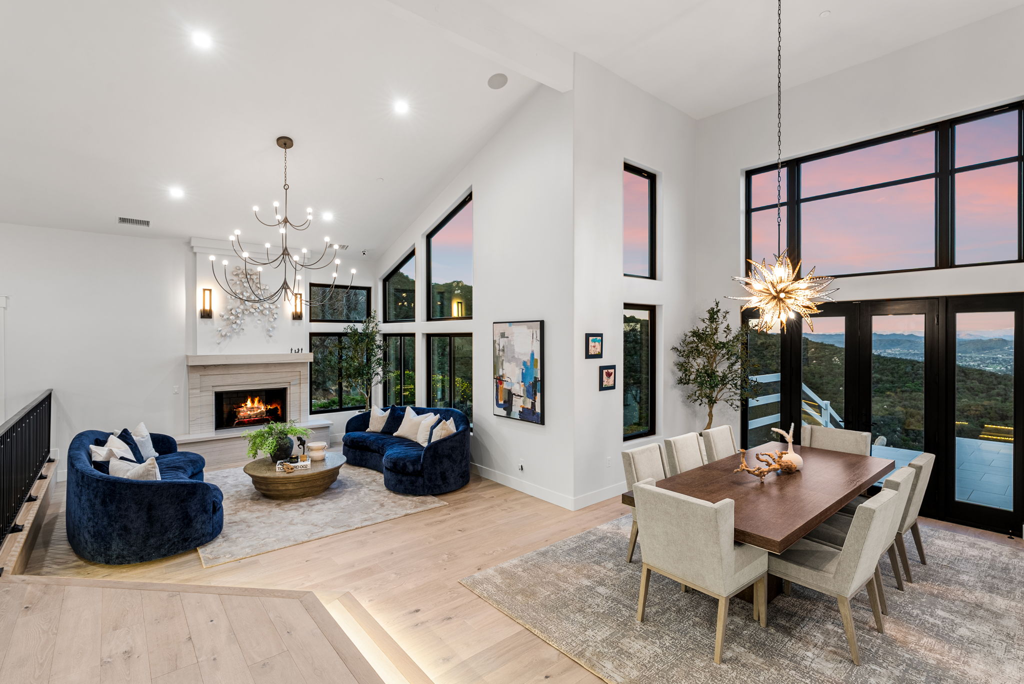 This stunning open-concept living and dining area features vaulted ceilings, light wood flooring, and floor-to-ceiling windows that capture sweeping mountain views. The space is anchored by a contemporary fireplace and furnished with plush, deep blue curved sofas, creating an elegant and airy atmosphere suitable for upscale entertaining. The integration of modern lighting fixtures, including a dramatic starburst chandelier, adds a touch of sophisticated luxury to this light-filled home.