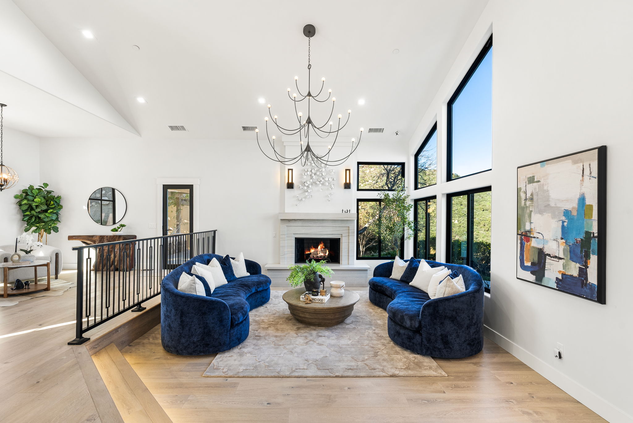 This sophisticated living room features a symmetrical layout with two plush navy blue curved sofas centered around a rustic wooden coffee table. A grand, modern chandelier acts as a focal point above a stone fireplace, while expansive windows and high vaulted ceilings enhance the sense of space and natural light. The room showcases a refined contemporary aesthetic with high-end finishes and an elegant, airy atmosphere.