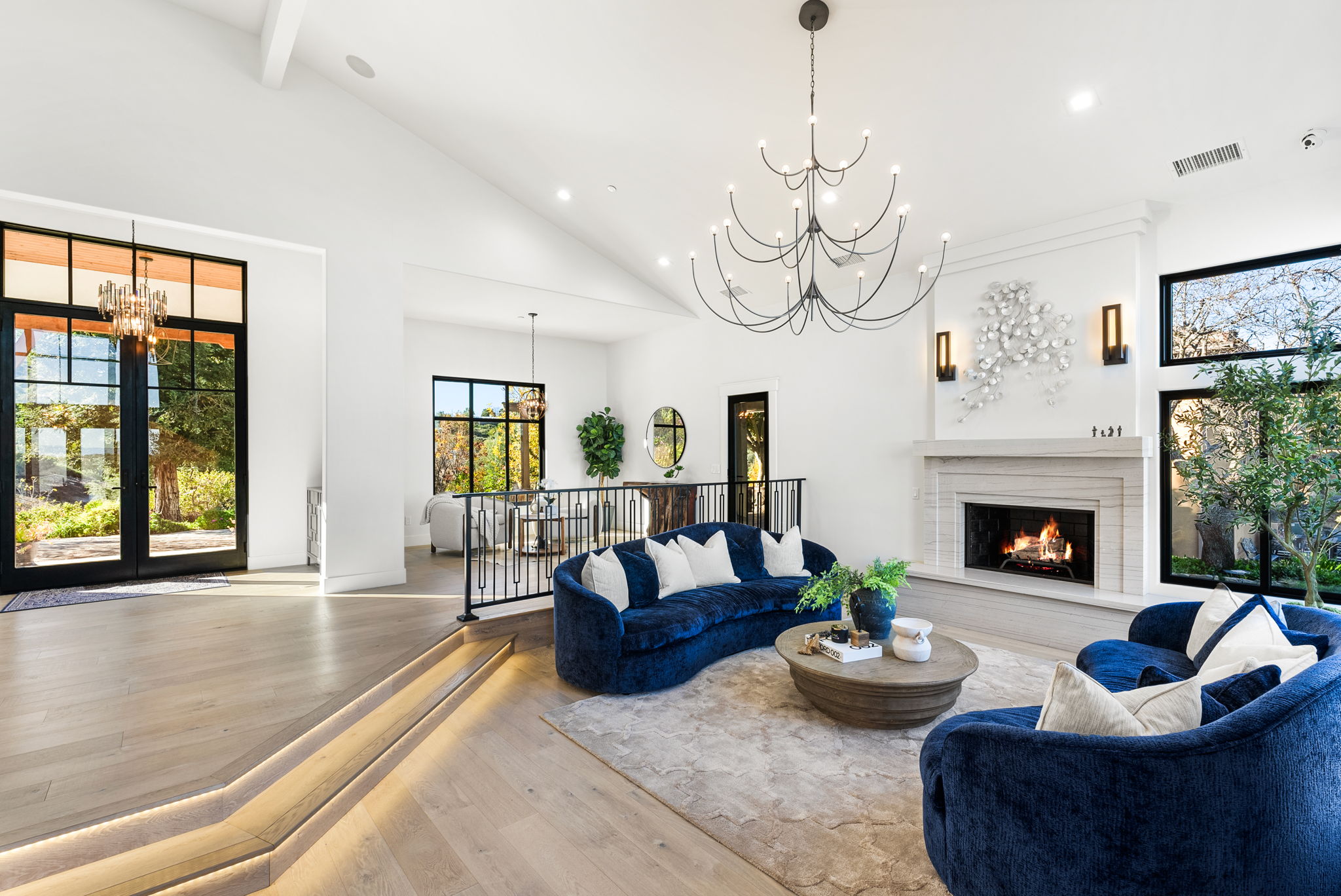 This spacious and elegant living room features a sunken floor design with illuminated steps, highlighting a luxurious blue velvet curved sofa set. The room is anchored by a grand stone fireplace with a decorative metallic art piece above and is bathed in natural light from large black-framed windows and doors. With its soaring vaulted ceiling, contemporary chandelier, and high-end finishes, the space exudes a sophisticated and modern atmosphere.
