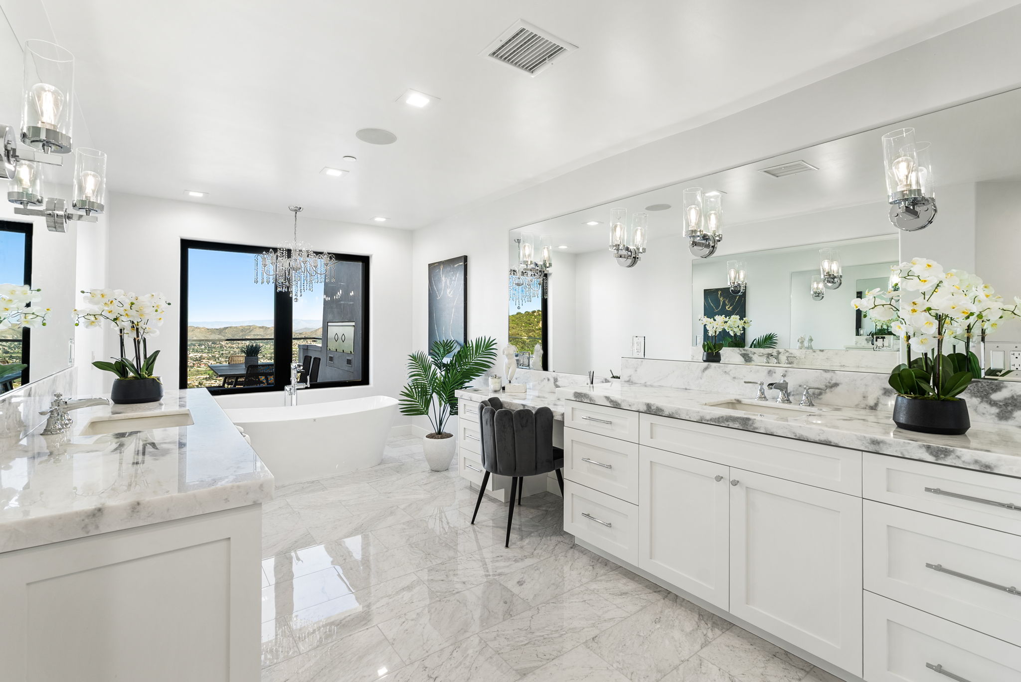 This luxurious primary bathroom features a bright, airy aesthetic with crisp white cabinetry, marble countertops, and expansive matching marble tile flooring. The room includes a standalone soaking tub situated by a large window with scenic views, a double vanity, and a dedicated vanity makeup station, all illuminated by sophisticated crystal lighting fixtures. The clean lines and high-end finishes create an elegant and spa-like atmosphere.