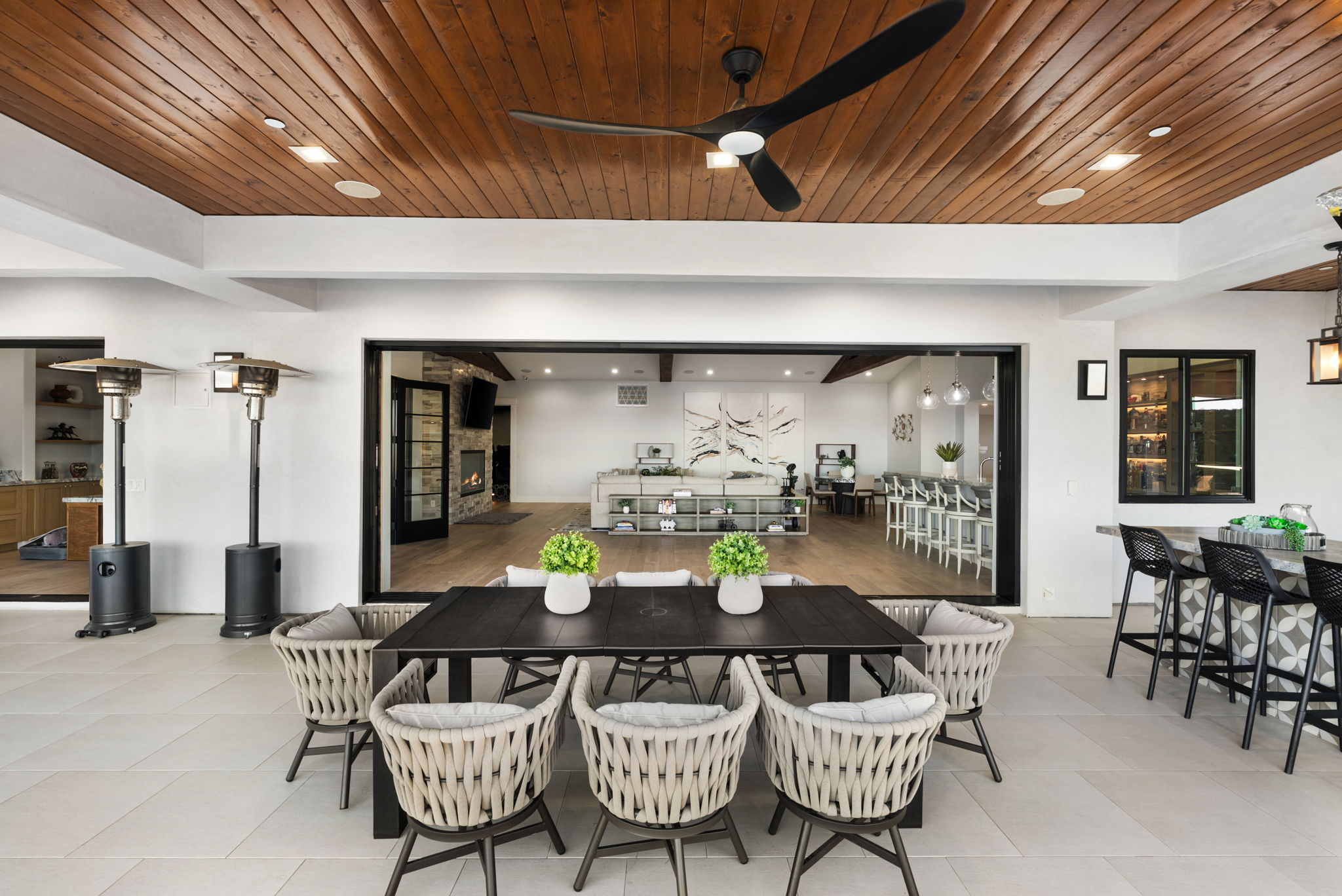 This spacious, covered outdoor patio features a large dining set with woven chairs and two decorative potted plants as a centerpiece. The space seamlessly integrates with the indoor living area through wide, retractable glass doors, showcasing a modern open-concept layout that includes a kitchen bar and living room. With high-end finishes, such as a wood-plank ceiling with a modern fan and recessed lighting, this area is perfect for luxury indoor-outdoor entertaining.