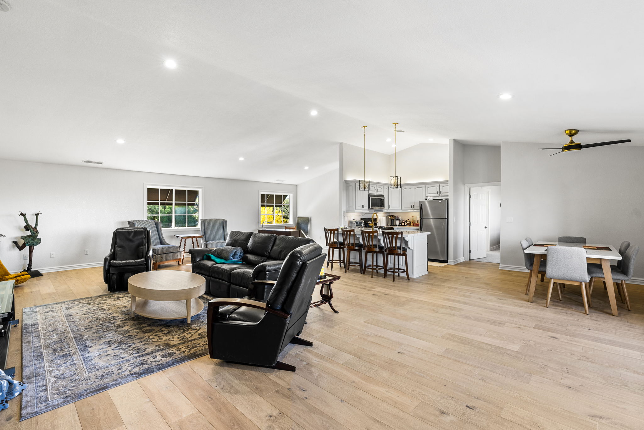 This open-concept living space features a spacious living room anchored by a large area rug and comfortable leather seating. The area flows seamlessly into a well-appointed kitchen with a breakfast bar, stainless steel appliances, and elegant pendant lighting. Large windows provide plenty of natural light, and the overall aesthetic is modern, bright, and inviting.