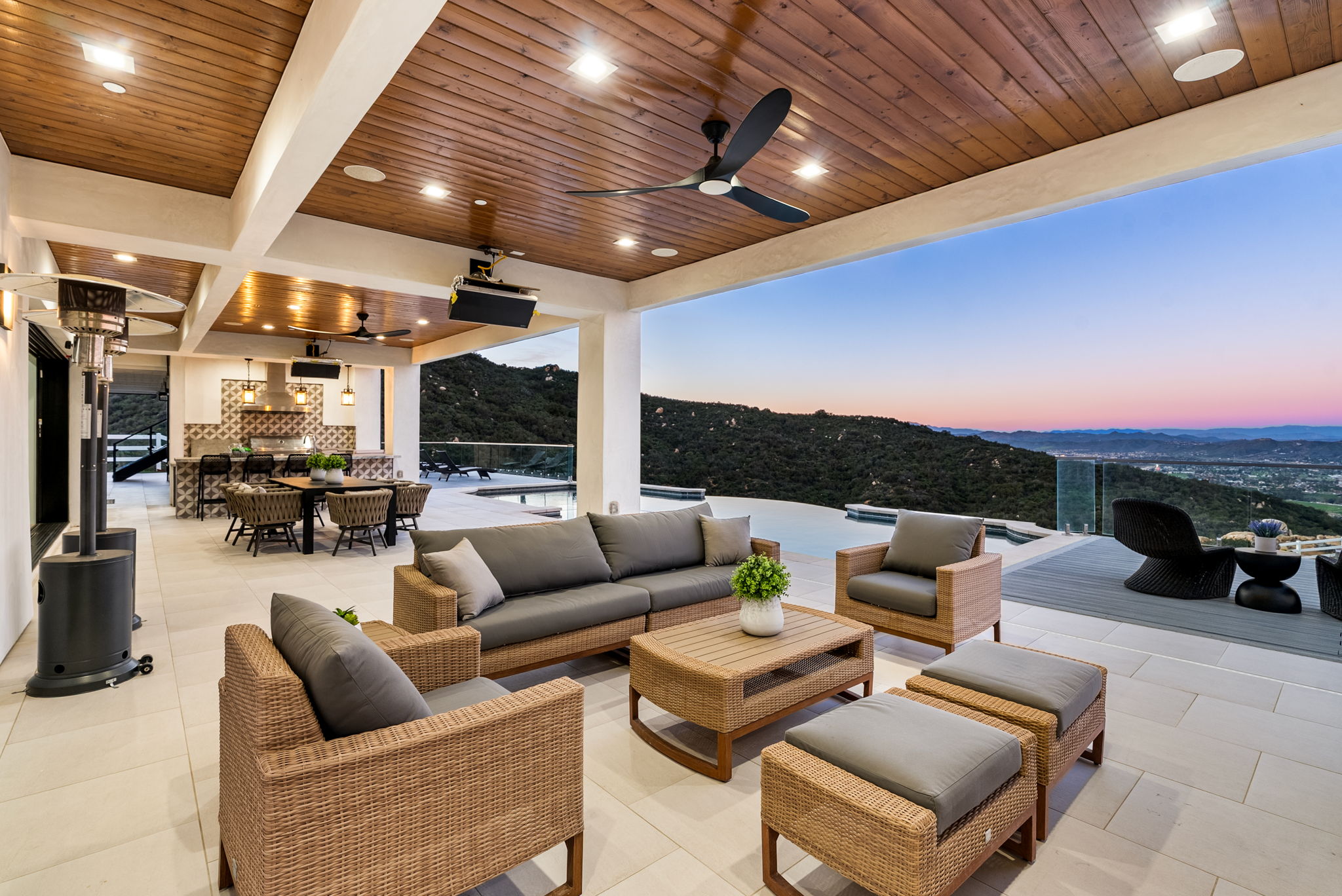 This luxurious covered outdoor living space features high-end wicker furniture, a cozy seating area, and a seamless transition to an infinity-edge pool overlooking expansive mountain views. The patio is elegantly designed with a wood-paneled ceiling, recessed lighting, a ceiling fan, and an adjacent full outdoor kitchen, making it perfect for refined entertaining. The overall atmosphere is sophisticated and highlights the property's stunning sunset vistas.