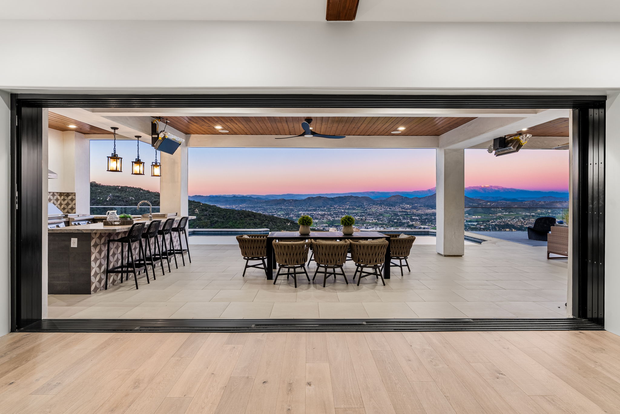 This stunning indoor-outdoor living space features a covered patio equipped with an outdoor kitchen, bar seating, and a formal dining area. The space seamlessly connects to the interior via large sliding glass doors and offers breathtaking, unobstructed panoramic views of the surrounding hills and city below. The modern design and high-end finishes create a luxurious atmosphere perfect for entertaining.