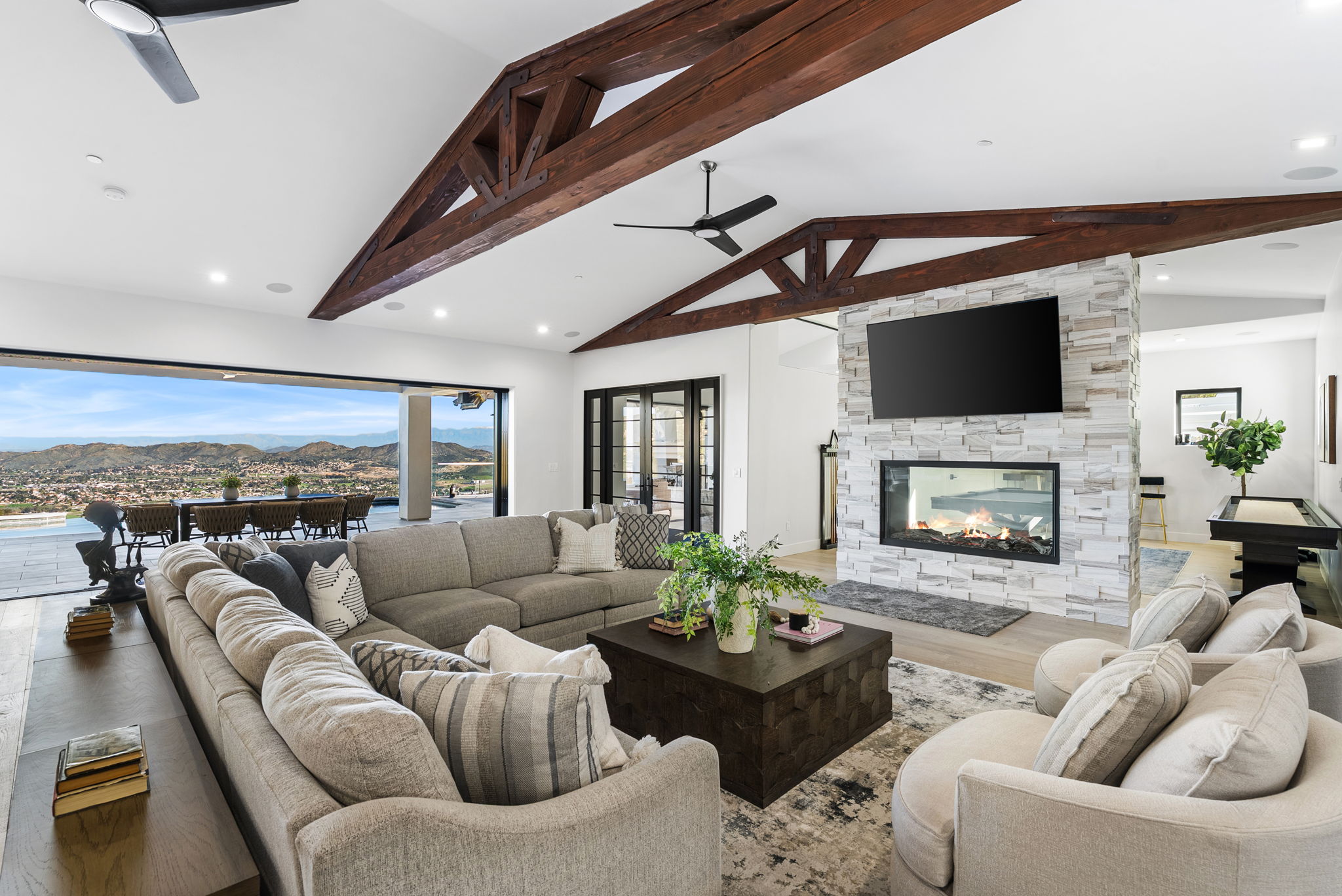 This spacious, open-concept living room features dramatic vaulted ceilings with exposed dark wood trusses and a grand stone fireplace with an integrated flat-screen TV. The room seamlessly connects to an expansive outdoor patio through sliding glass doors that showcase a stunning panoramic view of the surrounding landscape. Stylish, neutral-toned furniture and a central coffee table create a warm, inviting atmosphere perfect for both relaxing and entertaining.