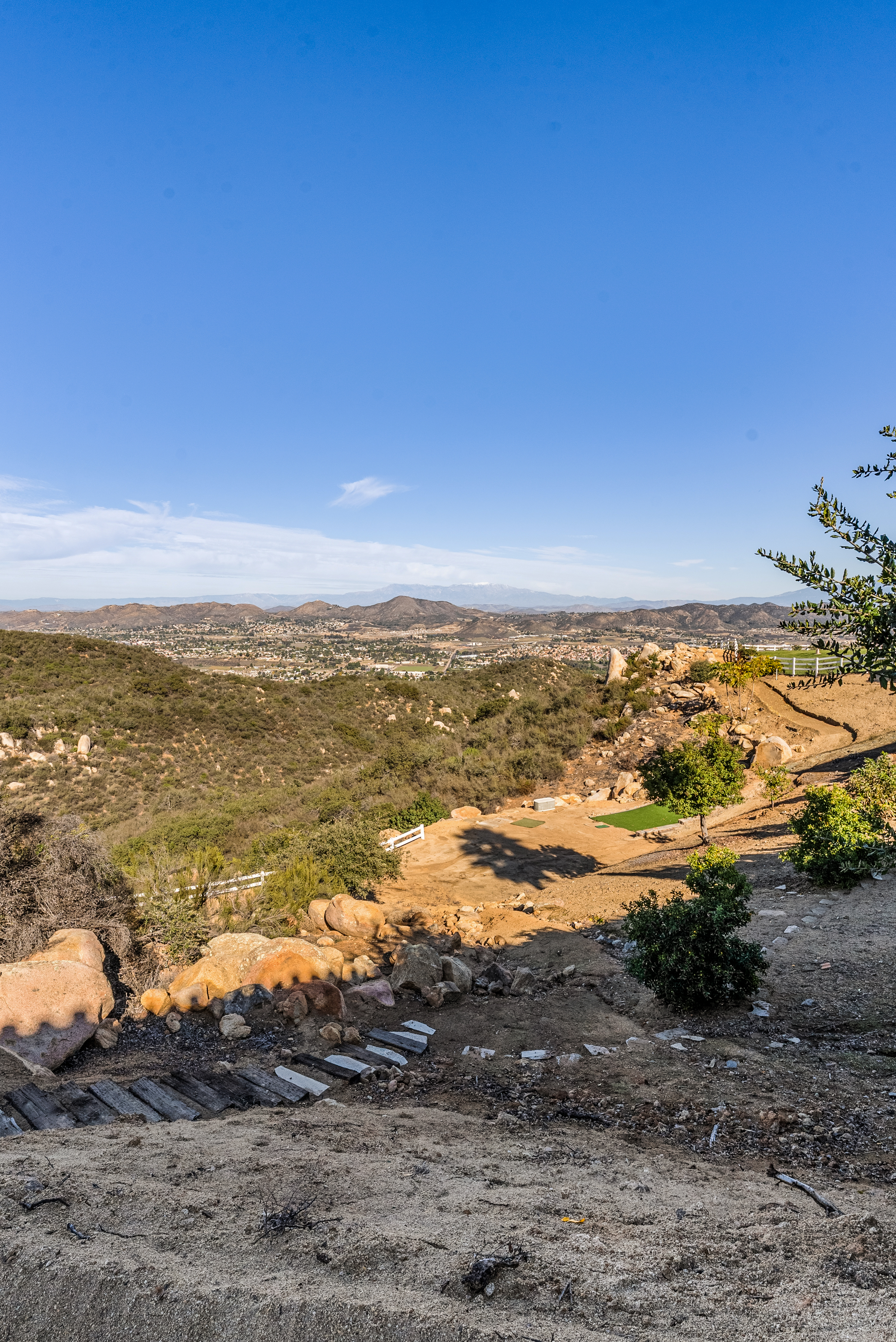 This elevated outdoor space offers sweeping panoramic views of the valley and distant mountains, suggesting a hillside property with natural landscaping. The area features a rustic path with wooden ties leading down to a tiered backyard section with patches of artificial turf, desert rocks, and small trees. It creates a serene, expansive environment ideal for enjoying natural scenery and outdoor relaxation.