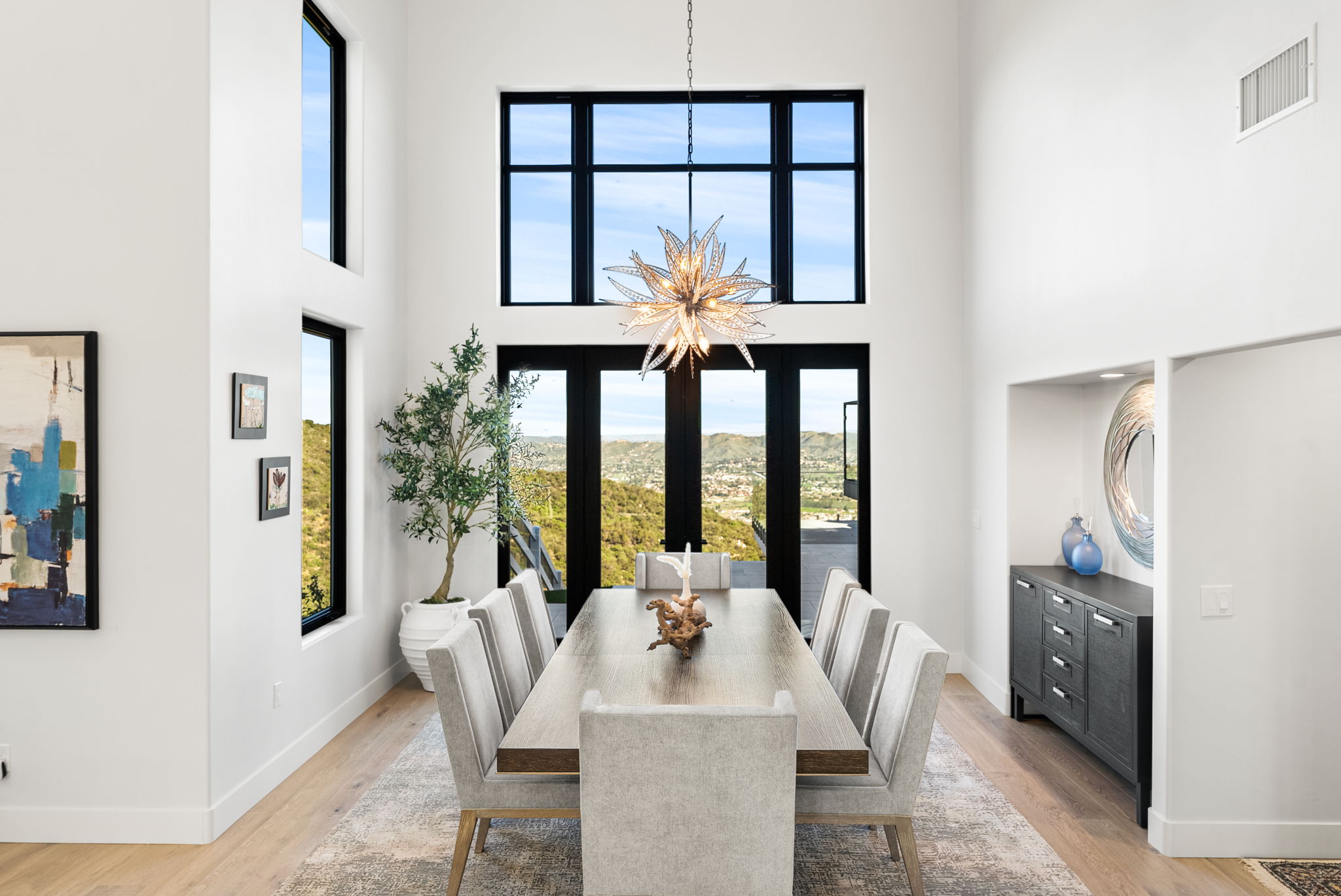 This sophisticated dining room features double-height ceilings and expansive floor-to-ceiling windows that offer panoramic hillside views. The space is anchored by a contemporary wooden dining table surrounded by eight upholstered chairs, all set beneath an artistic starburst-style chandelier. A dark wood sideboard and neutral-toned area rug complete the clean, modern aesthetic.