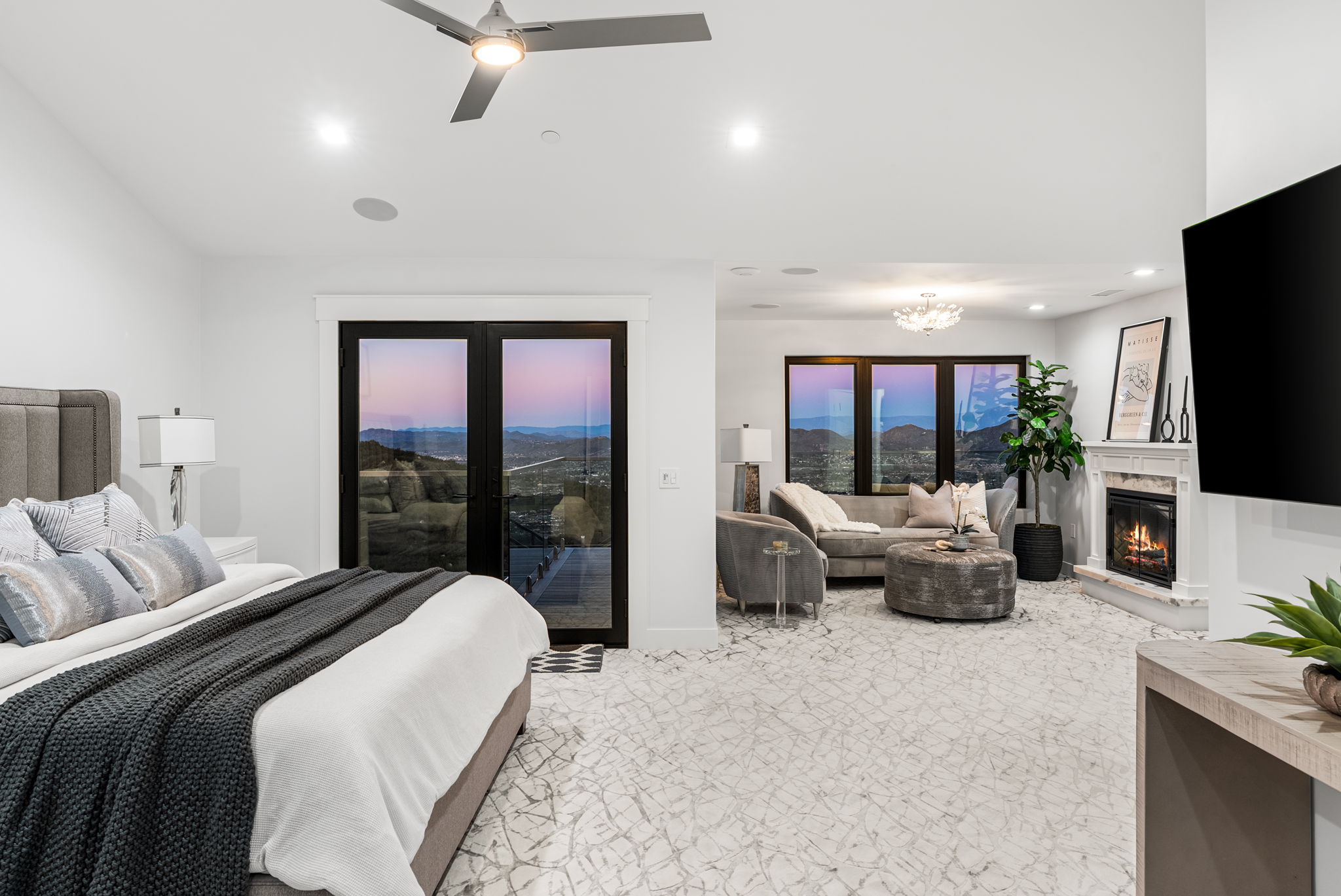 This spacious primary bedroom features a modern, high-end design with light marble-style flooring and an integrated sitting area complete with a fireplace. The room offers stunning sunset mountain views through large glass doors and windows, creating a serene and luxurious atmosphere. A large television, cozy seating, and a king-sized bed with textured linens make this suite both comfortable and sophisticated.