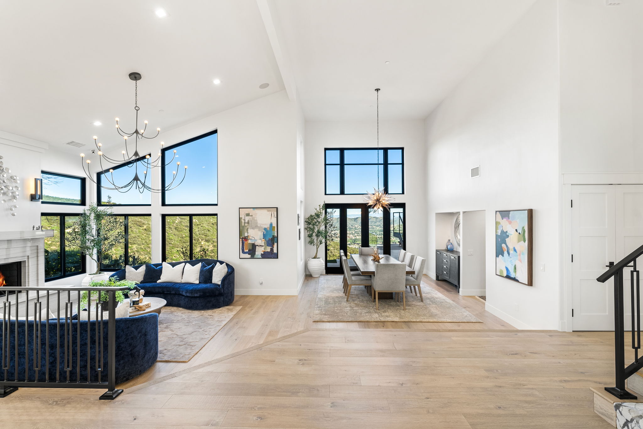 This spacious, open-concept living and dining area features high ceilings, light hardwood floors, and large windows that provide abundant natural light and scenic hillside views. The room is anchored by a sophisticated navy blue curved sofa in the living space and a modern dining set under a statement starburst chandelier. With its bright white walls, contemporary art, and clean aesthetic, the home offers an airy and elegant atmosphere suitable for both relaxation and entertaining.