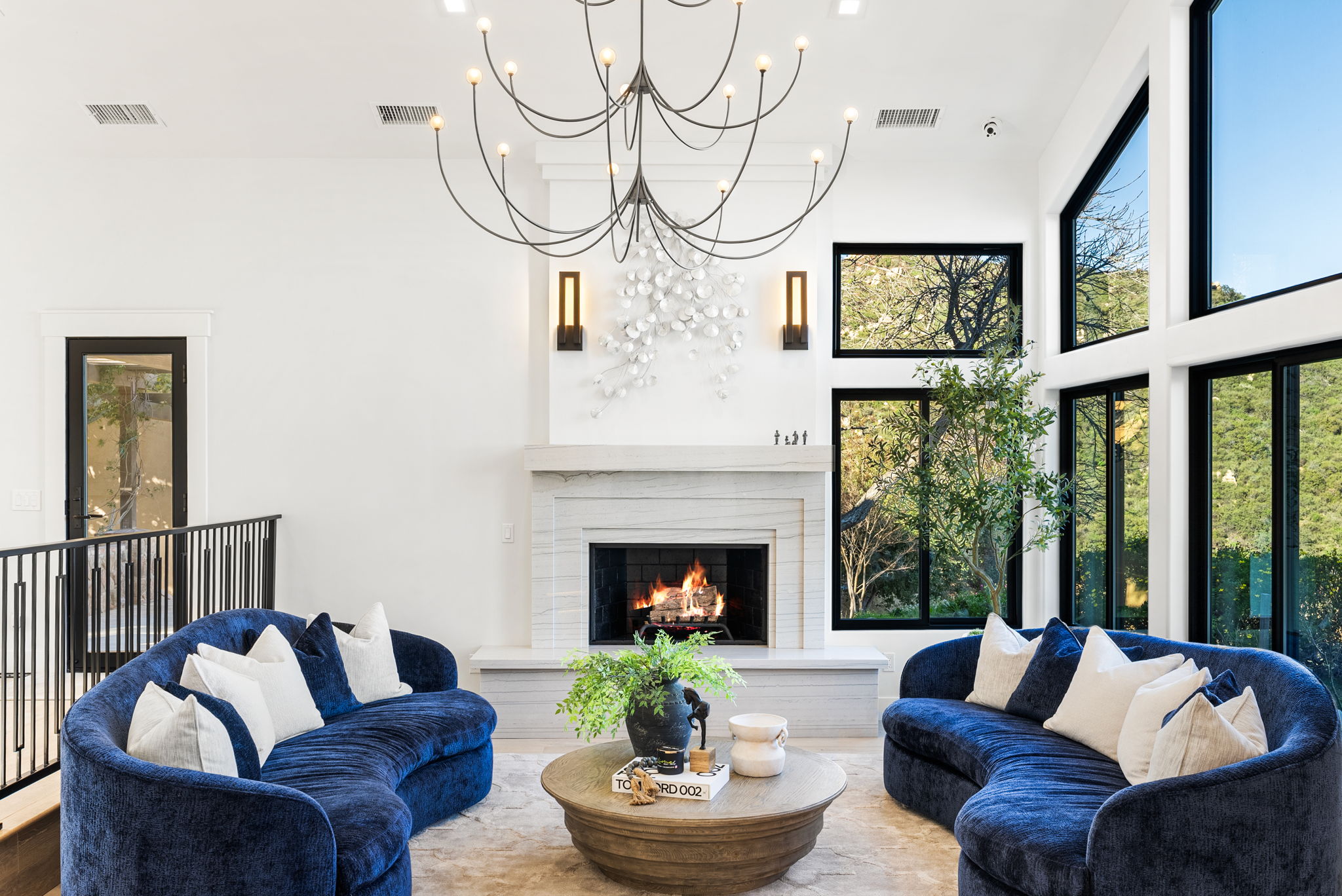 This elegant, high-ceilinged living room features two striking curved navy blue sofas arranged around a circular wooden coffee table, centered on a fireplace with a light stone surround. Large black-framed windows offer views of the outdoors, while a dramatic modern chandelier and wall sconces provide sophisticated lighting. The space exudes a luxurious, contemporary aesthetic with a clean and welcoming atmosphere.