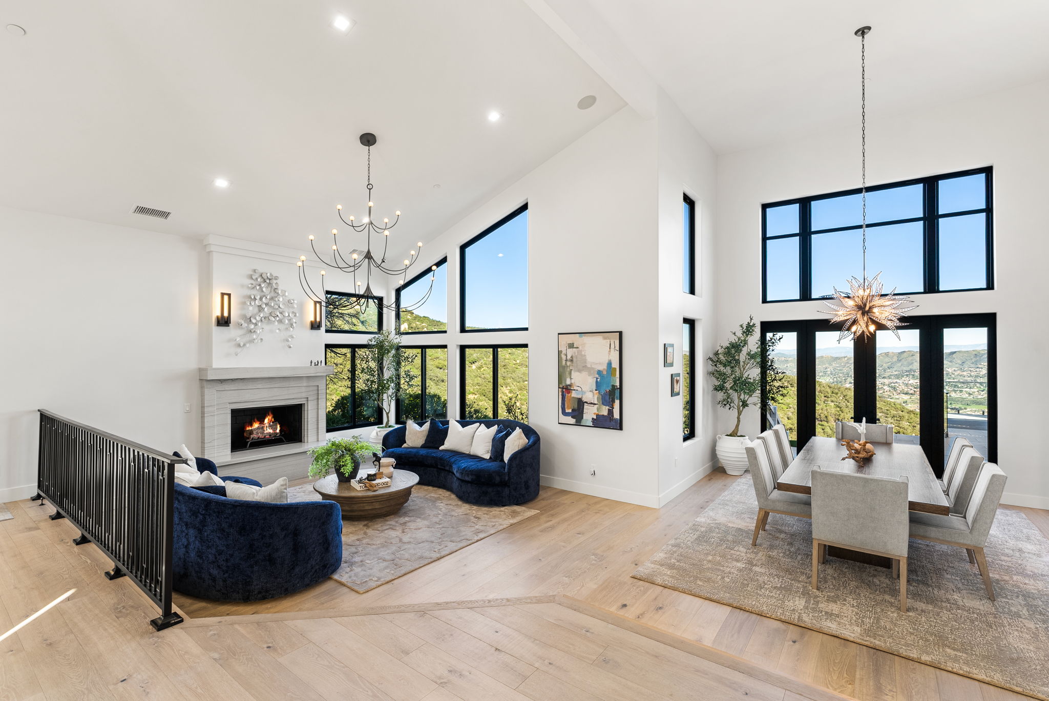 This stunning open-concept living area features soaring vaulted ceilings, a cozy fireplace with a modern mantle, and two deep blue curved velvet sofas. Large floor-to-ceiling windows flood the space with natural light while showcasing expansive hillside views, seamlessly integrating the sophisticated dining area adjacent to the living space. The interior is characterized by elegant modern lighting fixtures, light wood flooring, and a neutral color palette, creating a luxurious and welcoming atmosphere.