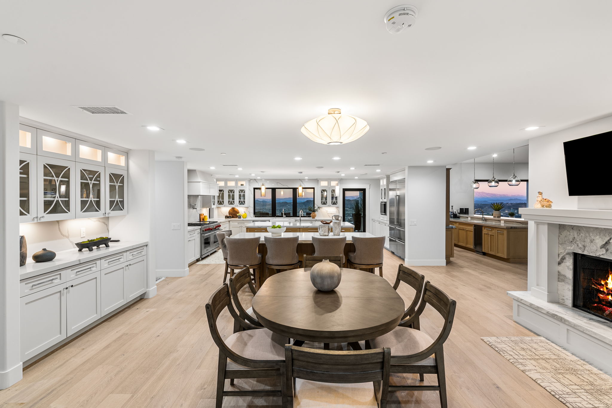 This spacious, open-concept kitchen seamlessly integrates dining and living areas, featuring a large central island with bar seating and light-toned hardwood flooring throughout. The space is anchored by a high-end gourmet kitchen with white cabinetry and a adjacent circular dining table, all complemented by a cozy fireplace and panoramic sunset views. It is a sophisticated, bright, and airy home design ideal for entertaining.