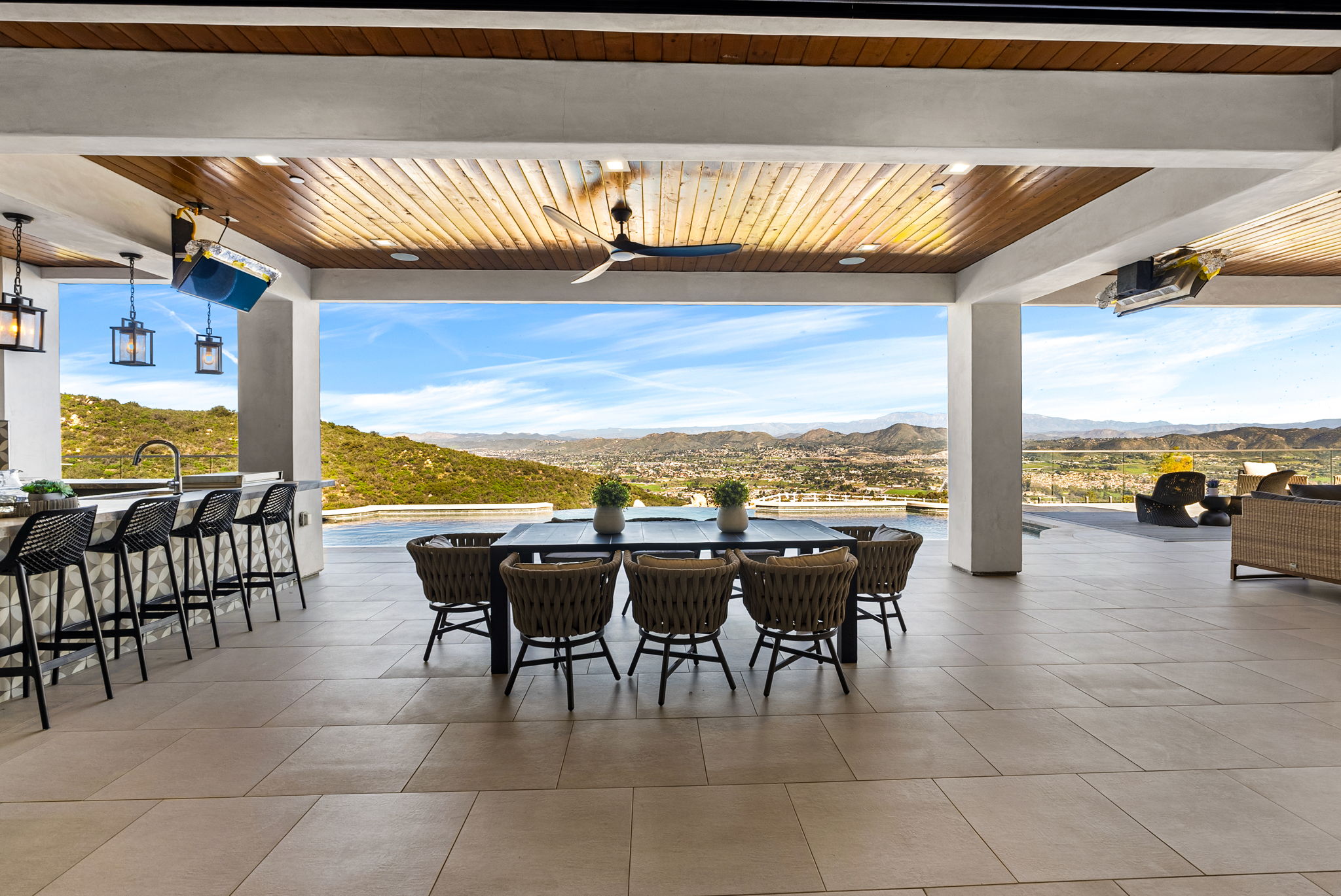 This spacious, covered outdoor patio offers a luxurious living and dining experience, complete with an integrated bar and a dining table that comfortably seats six. The area features high-end, woven furniture, modern lighting fixtures, and a stunning infinity-edge pool that seamlessly blends into the expansive panoramic mountain and valley views beyond. It is the perfect setting for upscale outdoor entertaining with a sophisticated, contemporary aesthetic.