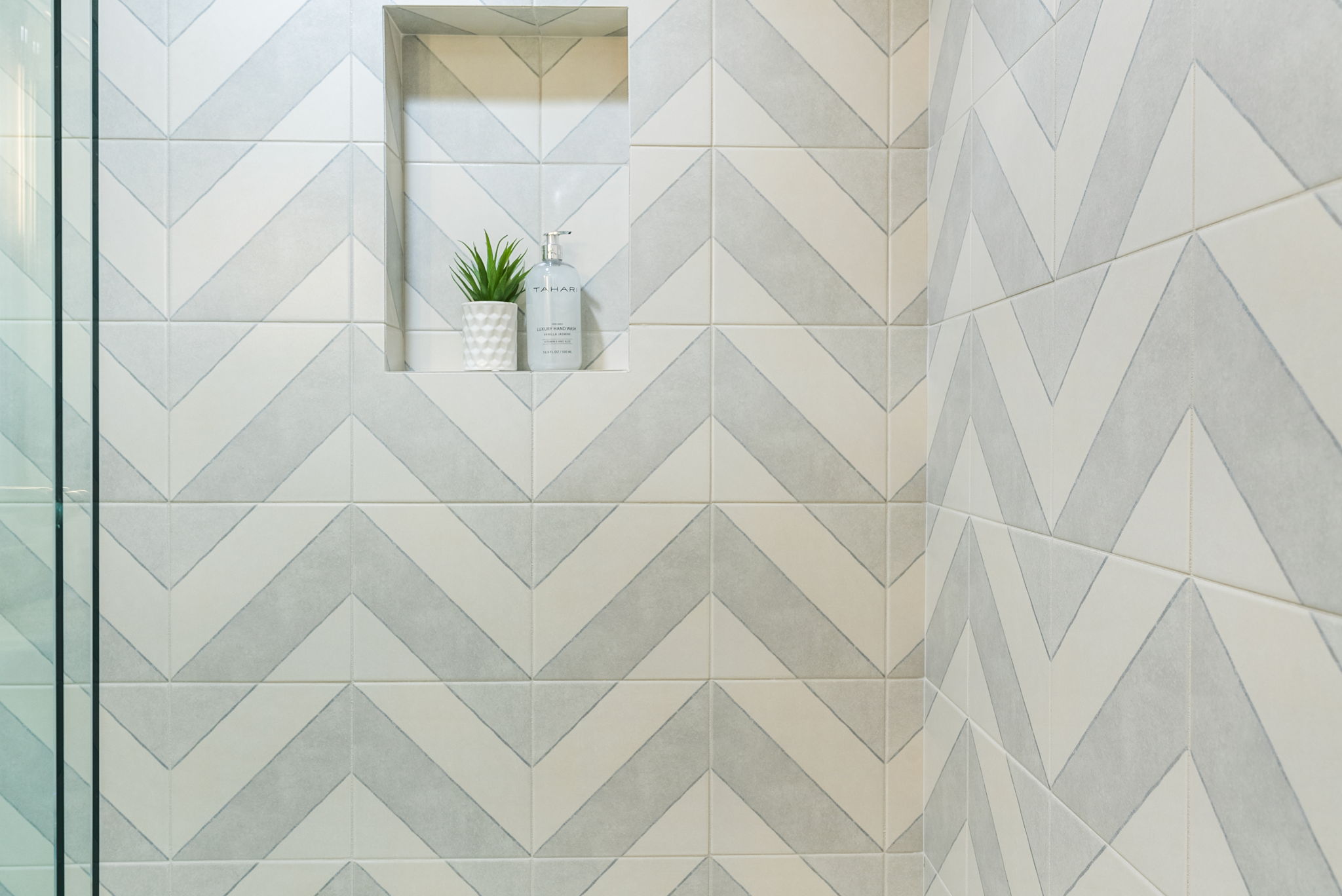 This image highlights a stylish shower wall featuring a chevron-patterned tile design in soft, neutral tones of cream and grey. A built-in recessed niche serves as a convenient storage area, currently holding a decorative potted plant and a luxury hand wash bottle, adding a functional yet elegant touch to the modern shower space.