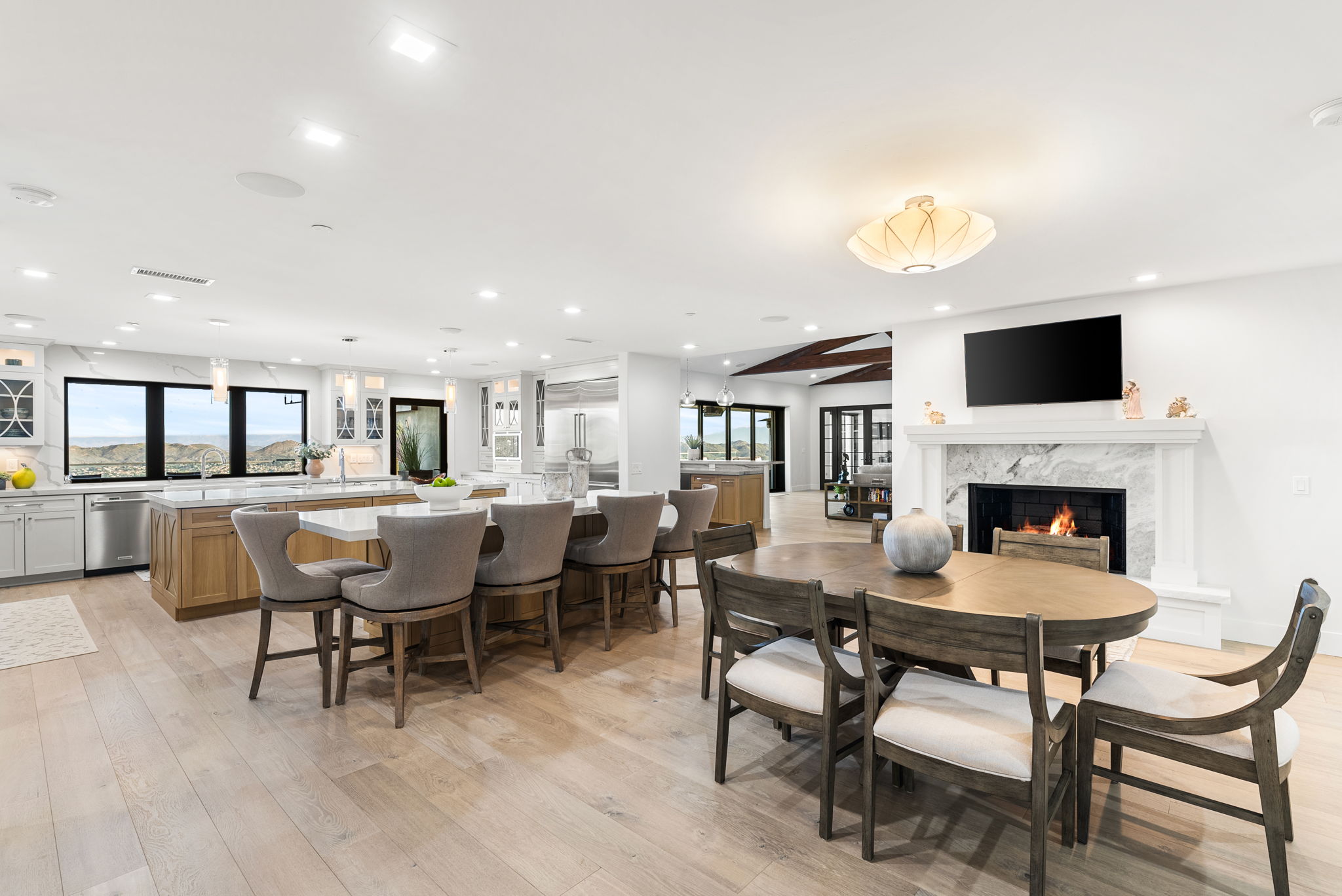 This spacious, open-concept kitchen seamlessly integrates with a formal dining area, featuring high-end cabinetry, a large waterfall-edge island with bar seating, and warm-toned wood flooring. The space is anchored by a cozy marble fireplace with an integrated television, creating an elegant blend of functional cooking area and comfortable living space. Bright and airy, the room is bathed in natural light from large windows that offer scenic mountain views, emphasizing a modern yet inviting design.
