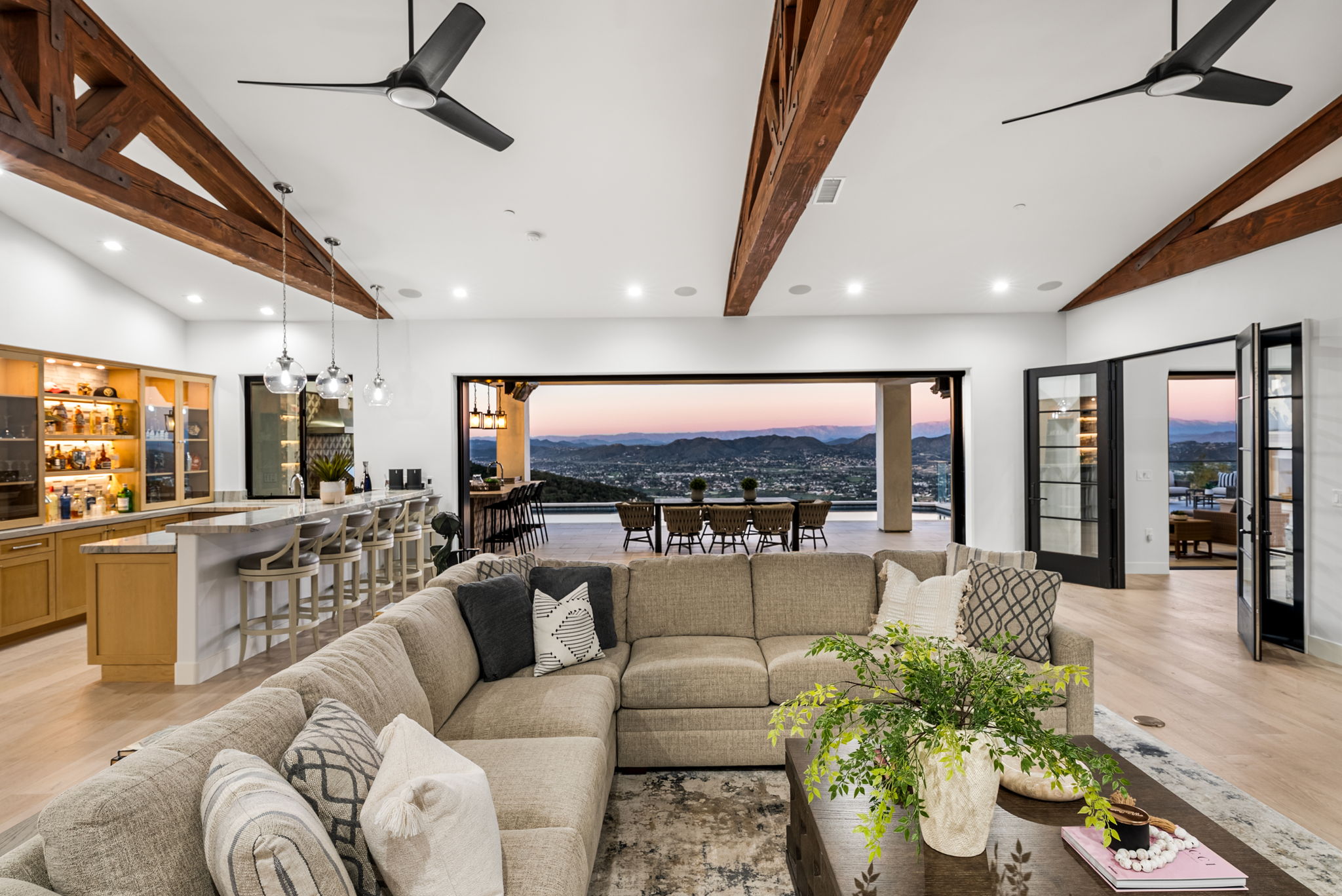 This spacious, open-concept living area seamlessly integrates with an outdoor patio, showcasing breathtaking panoramic mountain views through a massive retractable wall. The room features a cozy, large sectional sofa, rustic exposed wooden ceiling beams, and a well-appointed wet bar, creating an ideal setting for both relaxation and high-end entertaining. The sophisticated design balances contemporary comfort with warm, natural elements, highlighting the home's impressive architectural appeal.