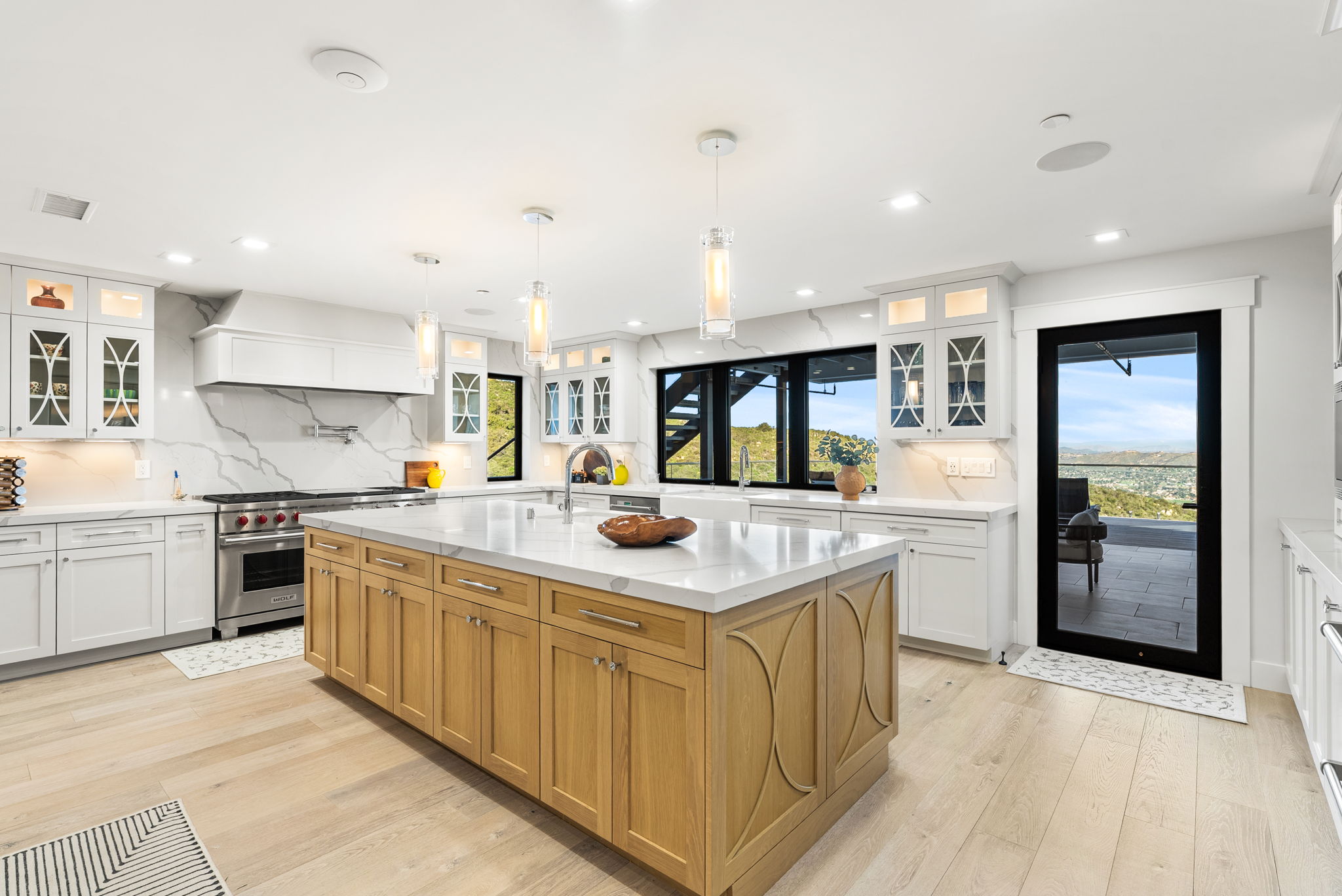 This bright and modern kitchen features a spacious light-oak kitchen island with a white marble-look countertop and matching cabinetry along the walls. The space is equipped with premium stainless steel appliances, including a Wolf range, and is illuminated by elegant glass pendant lights. Large windows and a glass door provide natural light and expansive outdoor views, creating a sophisticated and airy atmosphere.