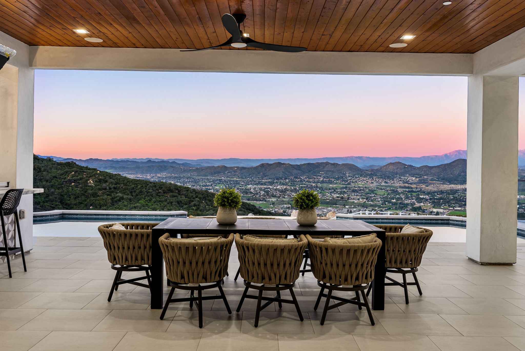 This stunning covered patio features a sophisticated dining area with a modern table and woven chairs, perfectly positioned to take advantage of breathtaking panoramic views. The space transitions seamlessly to an infinity-edge pool, overlooking a sweeping landscape of hills and a valley below at dusk. With a warm wood-paneled ceiling and recessed lighting, this outdoor area provides an exceptional, high-end environment for al fresco dining and relaxation.